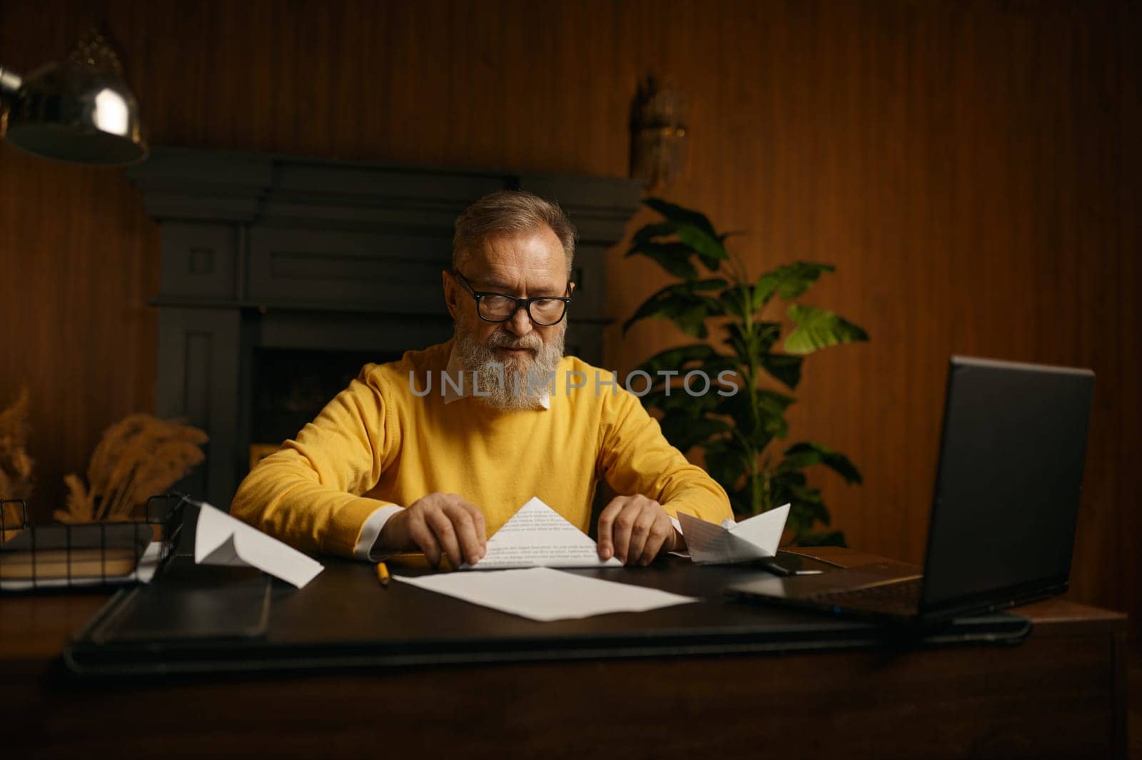 Bored senior man making paper origami figures while taking break or waiting for online conference videochat with business partners at home office workplace