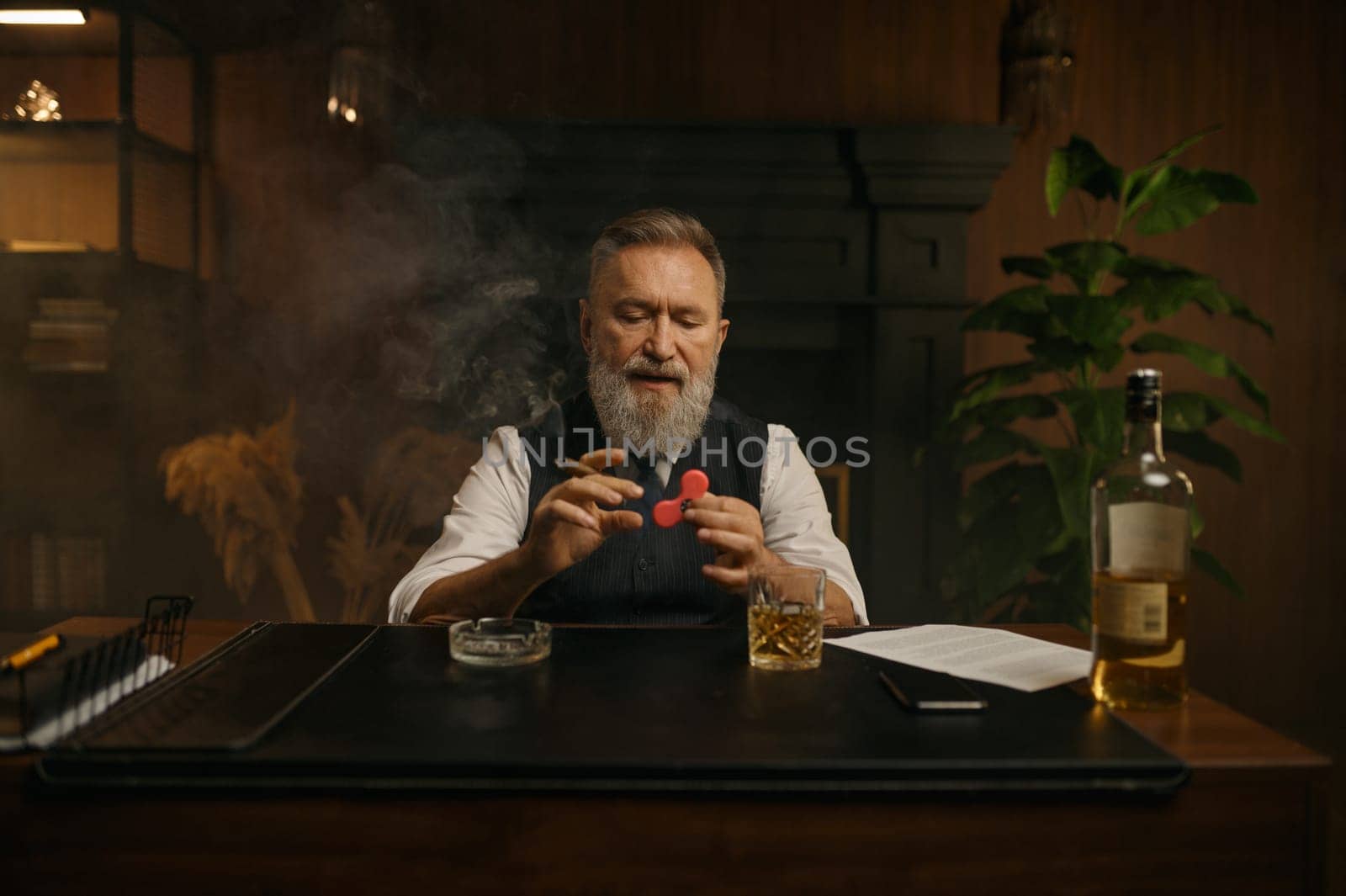 Old-fashioned elegant senior businessman playing spinner toy while sitting at work table over home office background