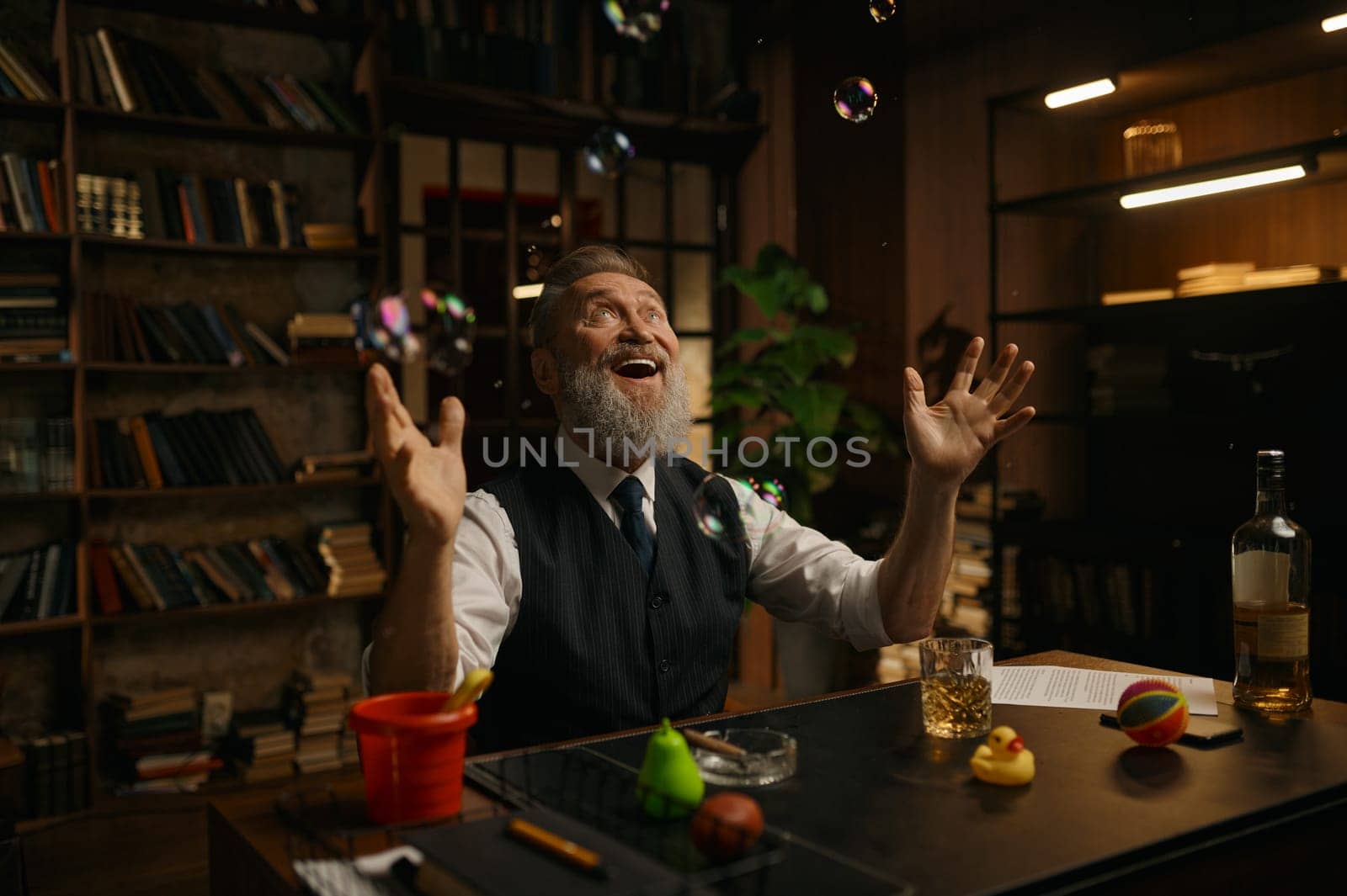 Happy overjoyed and playful funny senior businessman sitting at desk table under bubble blower rejoicing and feeling joy