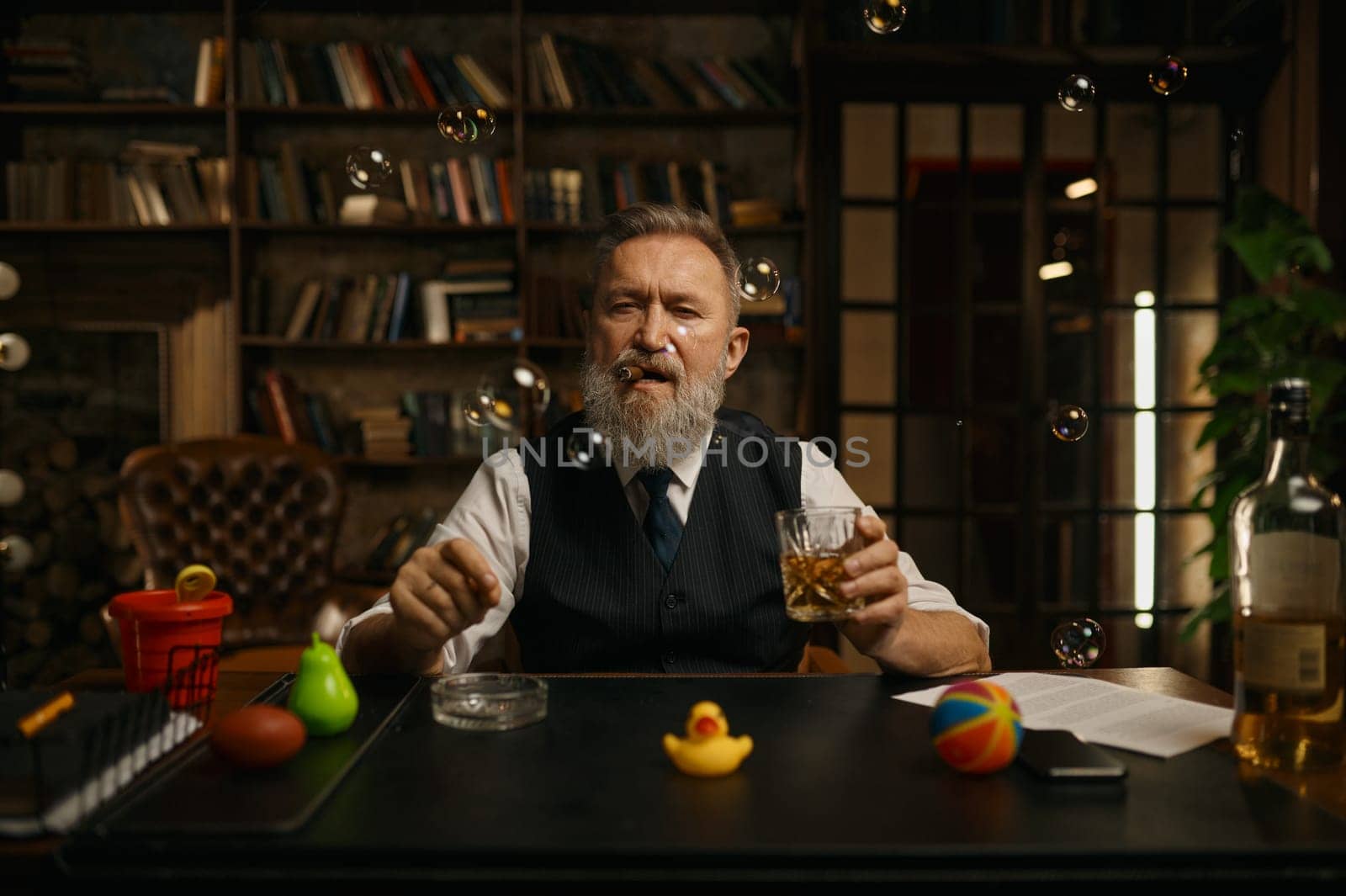 Portrait of relaxed senior businessman smoking cigar and drinking whiskey while sitting at work table in home office. Looking at camera under bubble blower flying in air