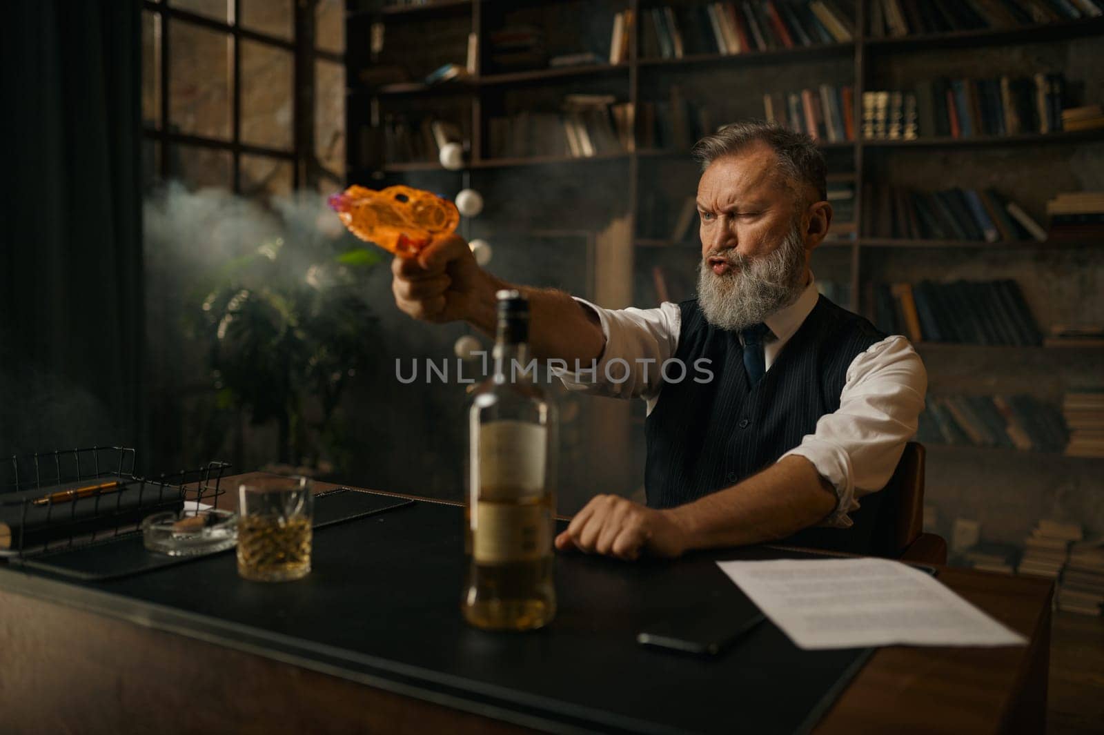 Brutal senior man shooting toy gun while sitting at table with alcohol drink and paper contract agreement document. Celebrating of good deal and overcoming competitors concept