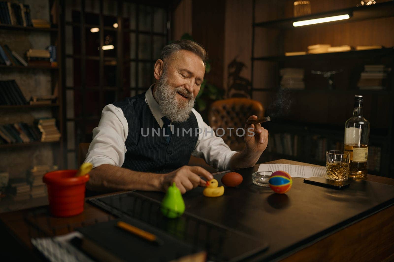 Funny elegant older businessman smoking cigar and playing toys at work table in home office