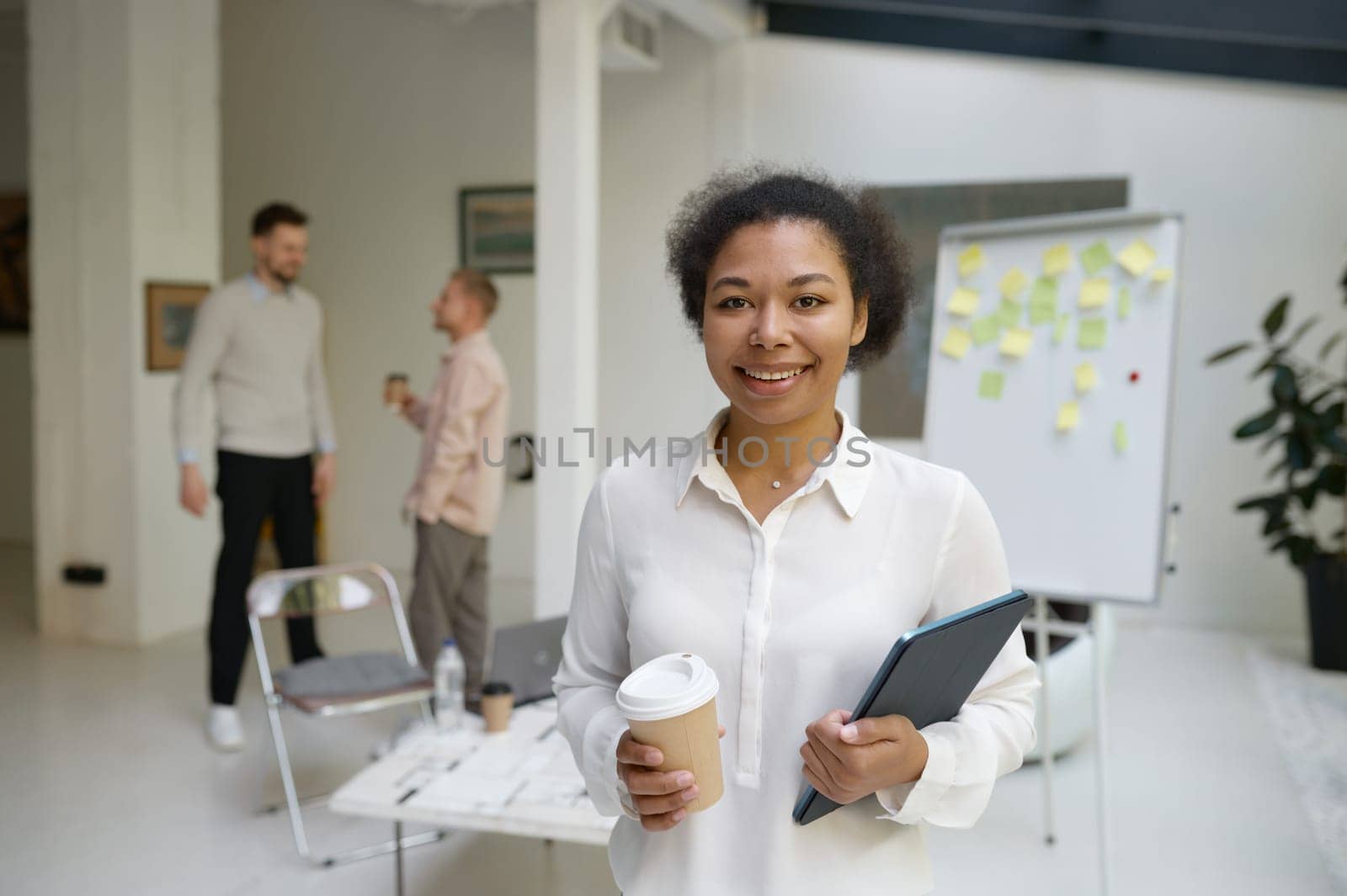 Portrait of female office worker holding digital tablet in hand looking forward while business colleagues talking on blurred background. Successful meeting and negotiation concept