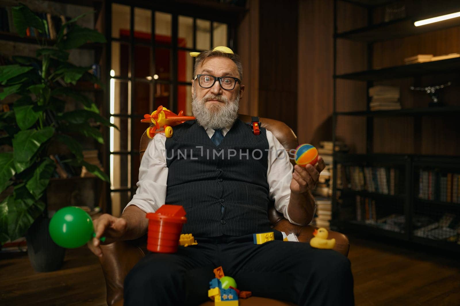 Portrait of rich senior businessman sitting in leather armchair with toys looking at camera over cozy home office interior