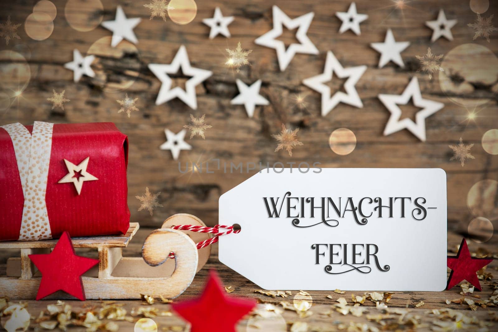 Wooden Sled With Red Gift And Rustic Christmas Bokeh Background With Label With German Text Weihnachtsfeier, Which Means Christmas Party In English