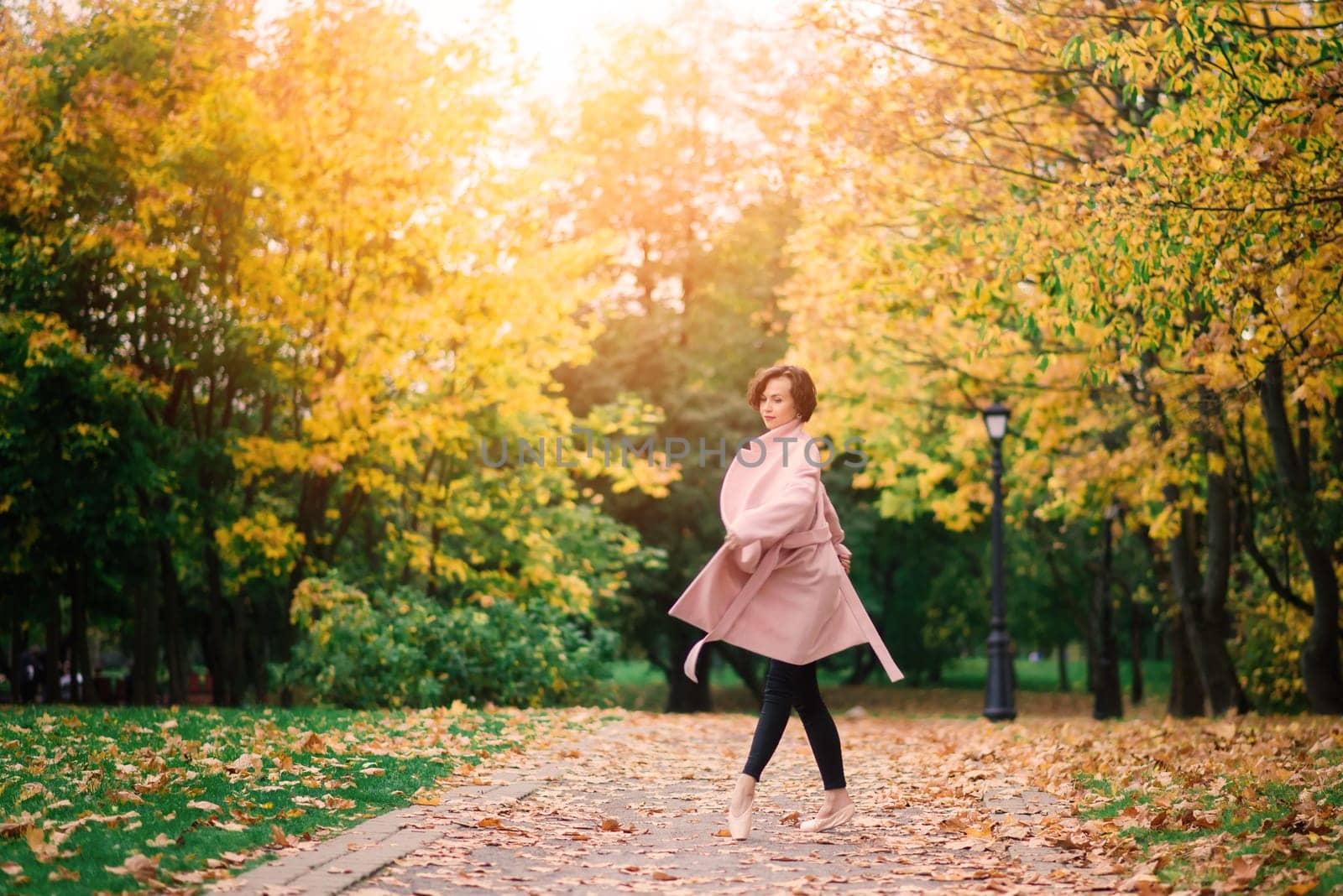 Woman ballerina dancing in pointe shoes in golden autumn park, standing in ballet pose on yellow leaves