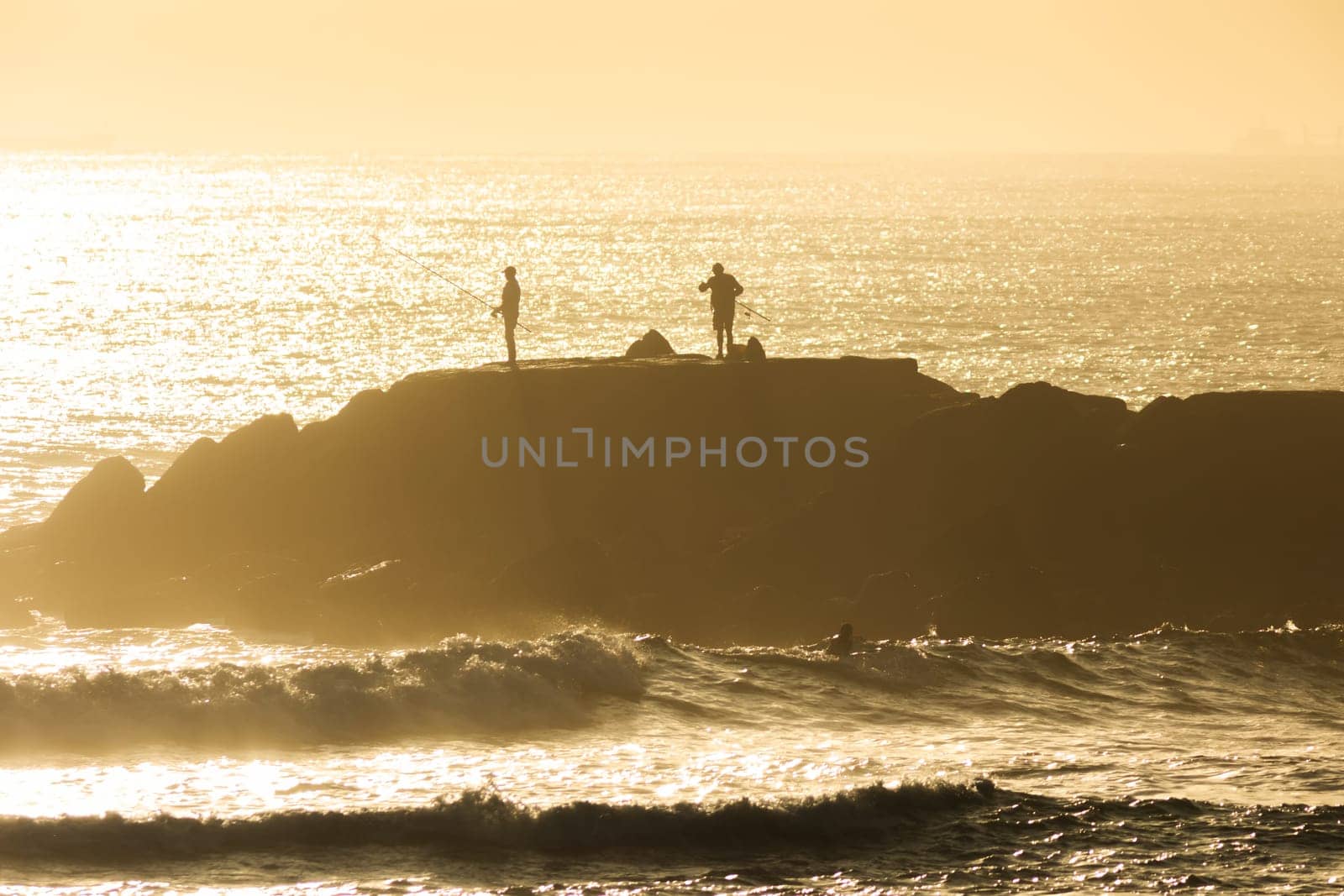 Silhouettes of people standing on a cliff in the middle of the sea and fishing at sunset. Mid shot