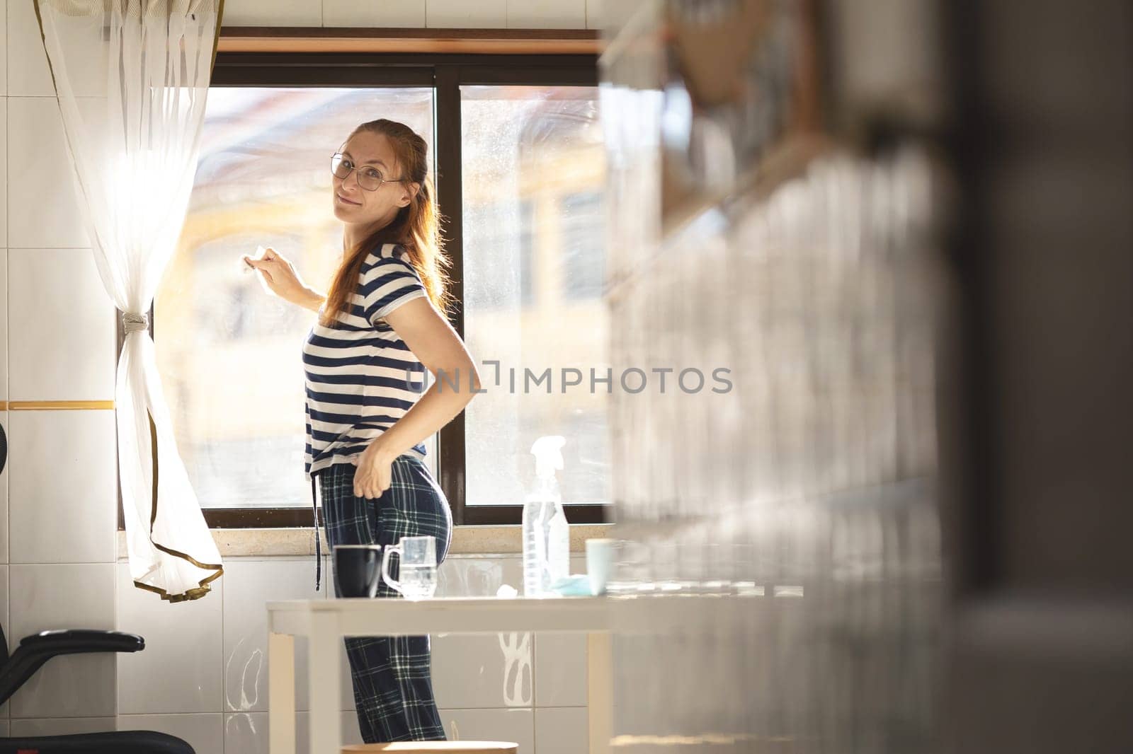 Adult woman housewife washing a window at the kitchen - looking in the camera and smiling. Mid shot
