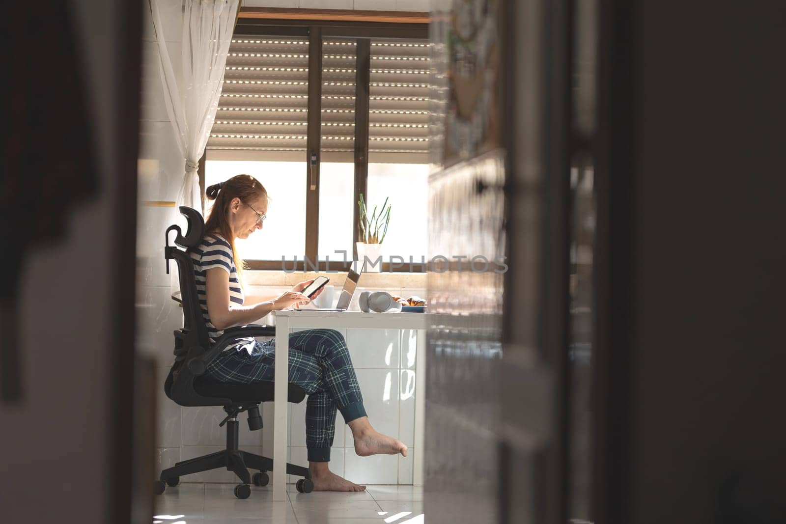 An adult woman working at her laptop at the kitchen table holding a phone. Mid shot