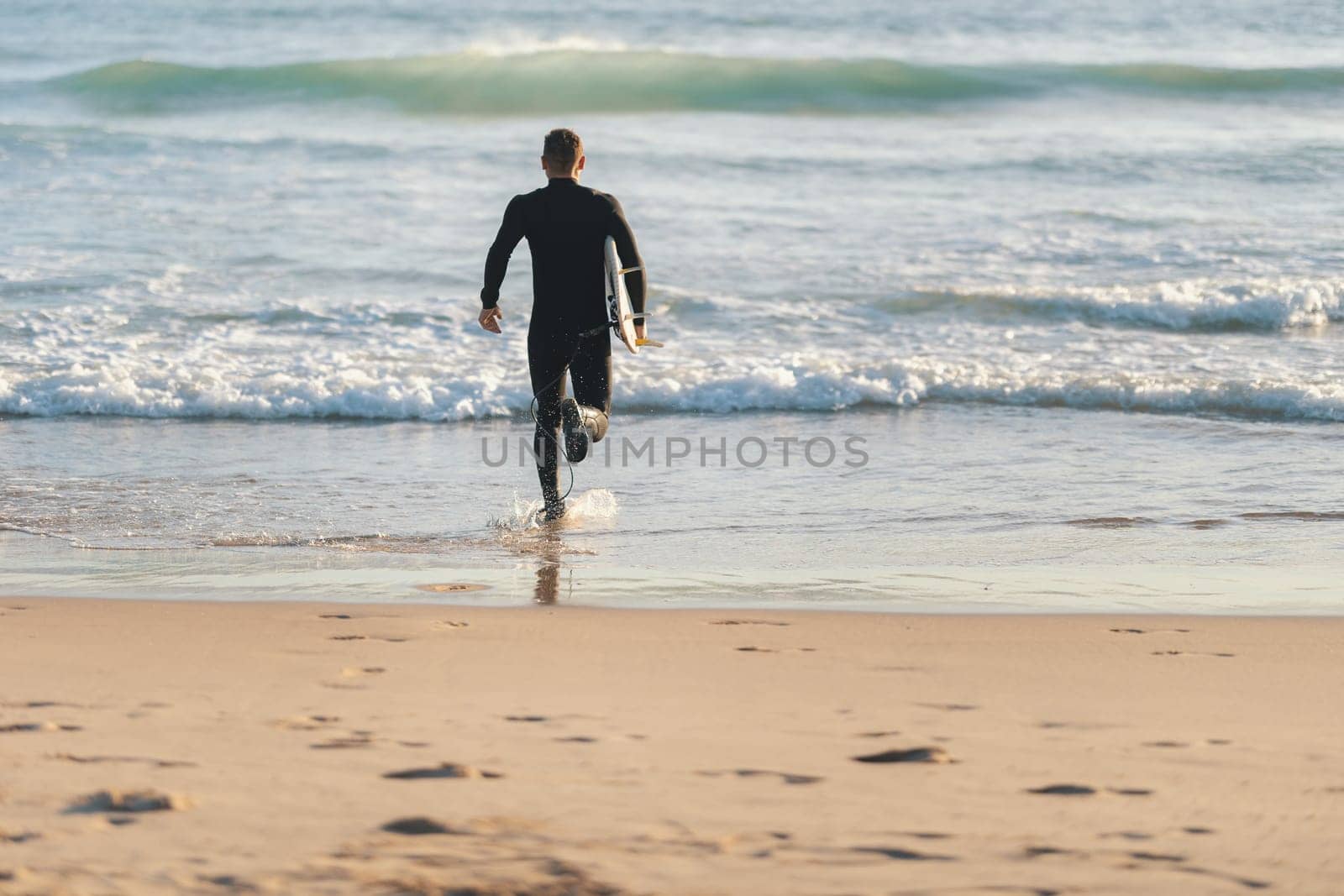 An athletic man surfer in a wetsuit running to the ocean holding a surfing board. Mid shot
