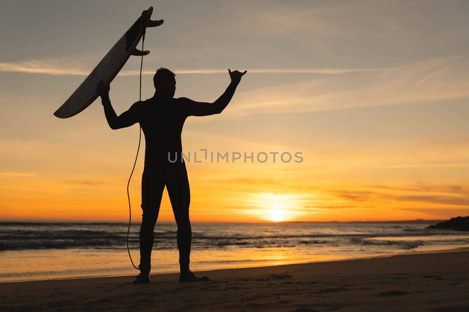 Silhouette of an athletic man standing on the seashore holding a surfboard and showing shaka at sunset. Mid shot