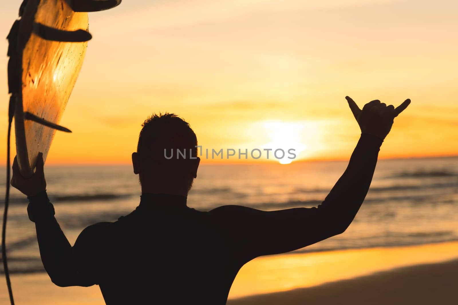 Silhouette of a man holding a surfboard showing shaka at sunset. Mid shot