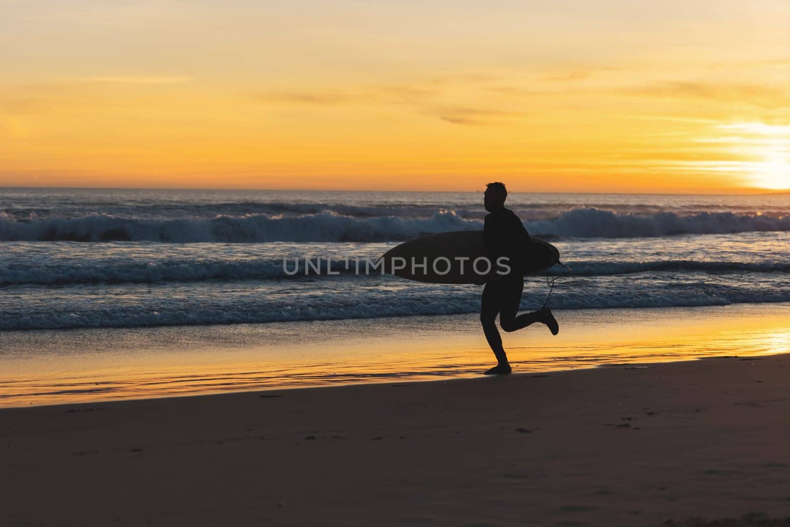 A man surfer running on the seashore holding a surfboard at sunset. Mid shot