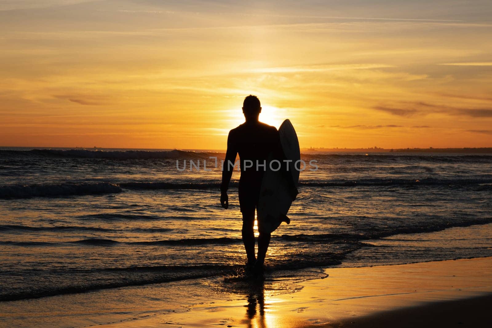 Dark silhouette of a man surfer walking out on the seashore holding a surfboard at sunset. Mid shot