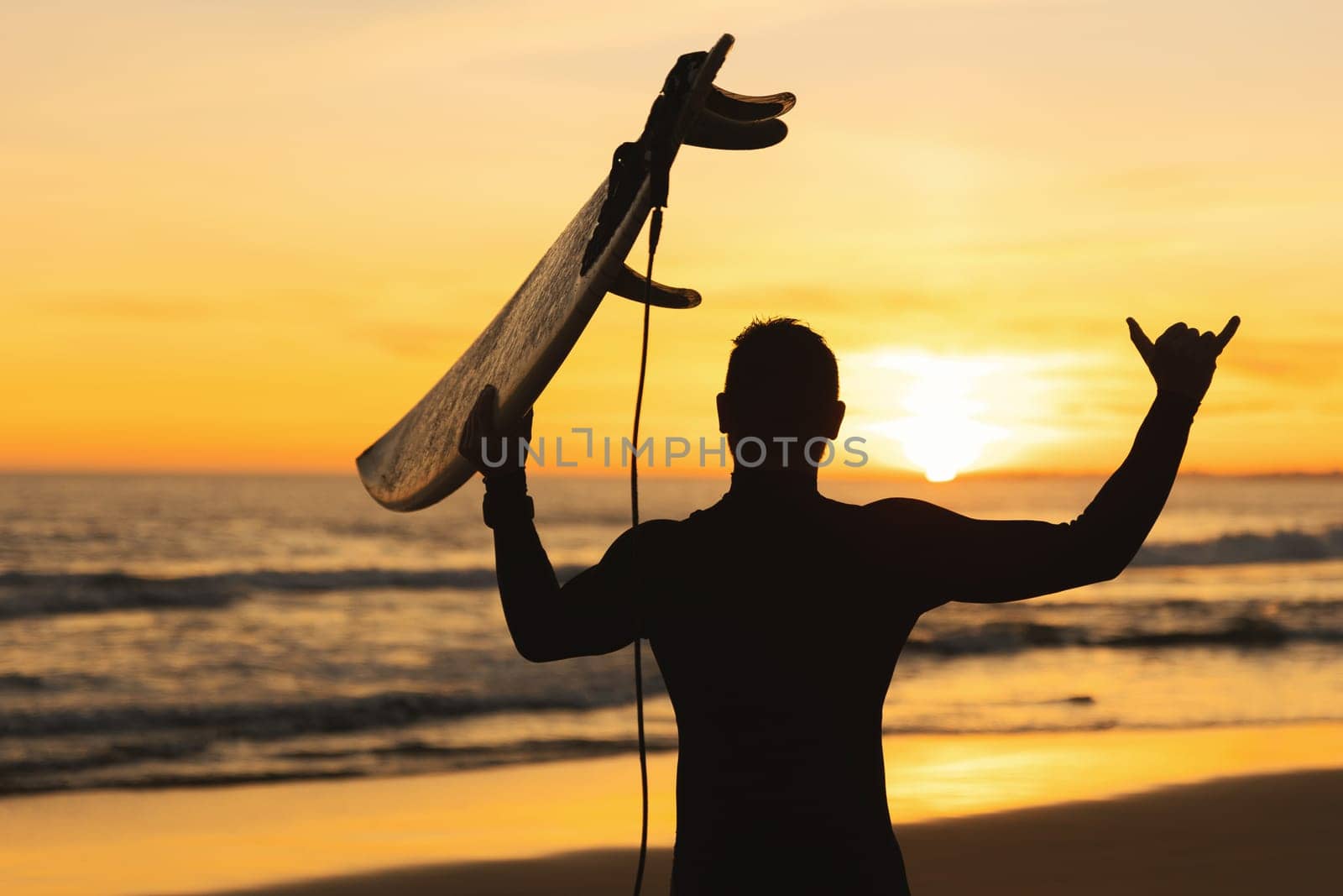 Silhouette of an athletic man holding a surfboard showing shaka at sunset. Mid shot