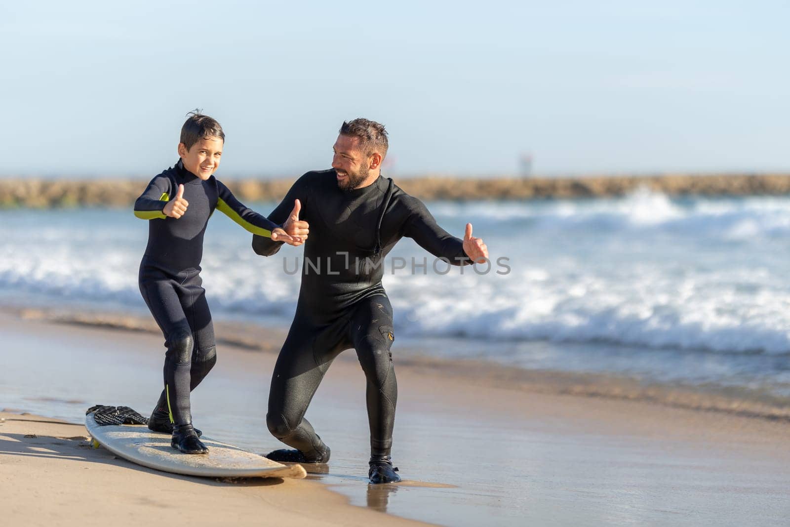A little boy stands on the surfboard on the seashore and his father standing by him - showing thumbs up. Mid shot