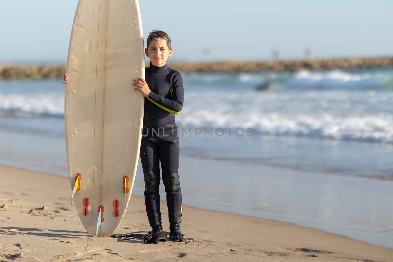 A little boy standing on the seashore with a big surfing board. Mid shot