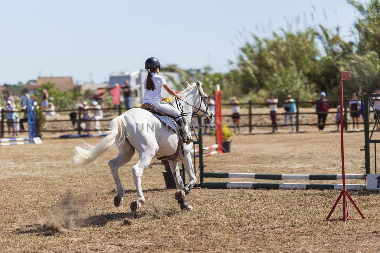 Equestrian sport - a little girl in uniform riding white horse at the ranch - jumping over the obsticles. Mid shot