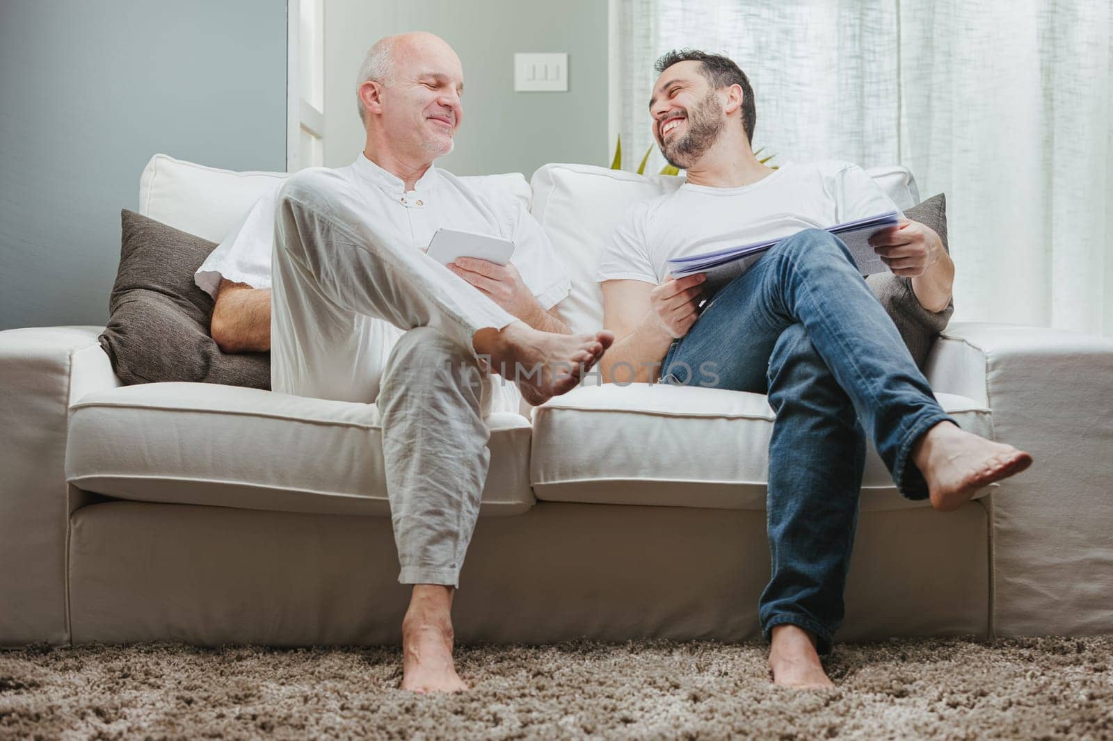 Father and son or couple of men relaxing together barefoot on a sofa laughing and smiling as they chat
