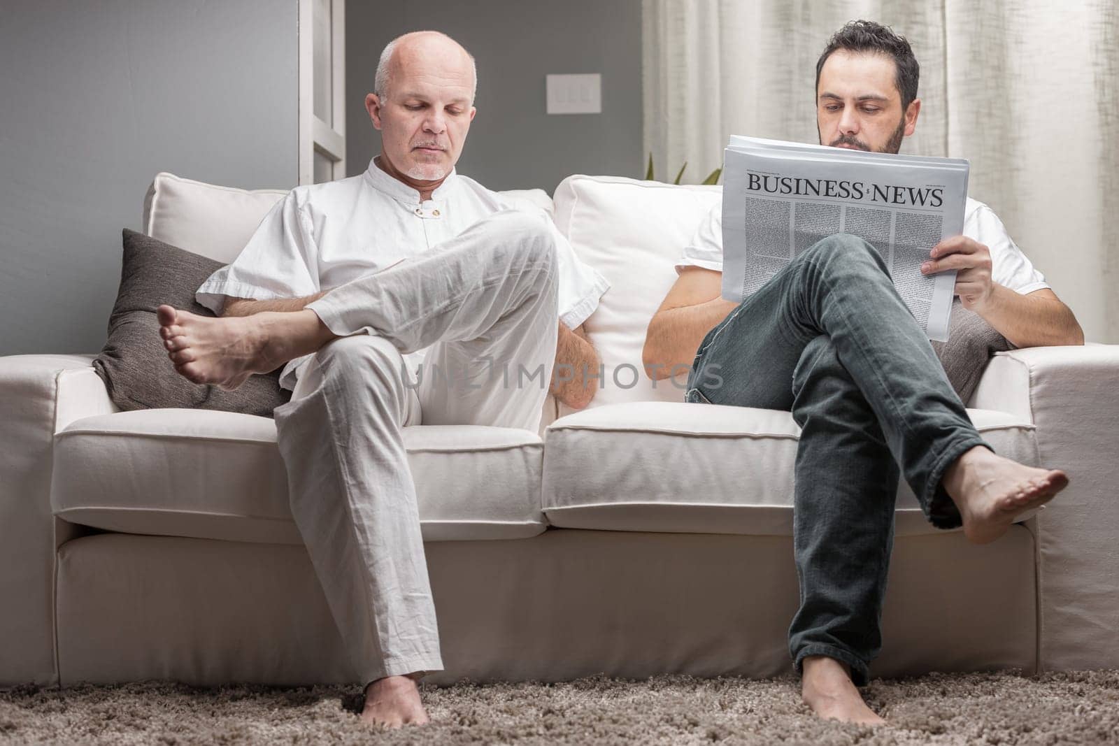 Father and son reading business news from a printed newspaper while sitting on a comfortable couch at home