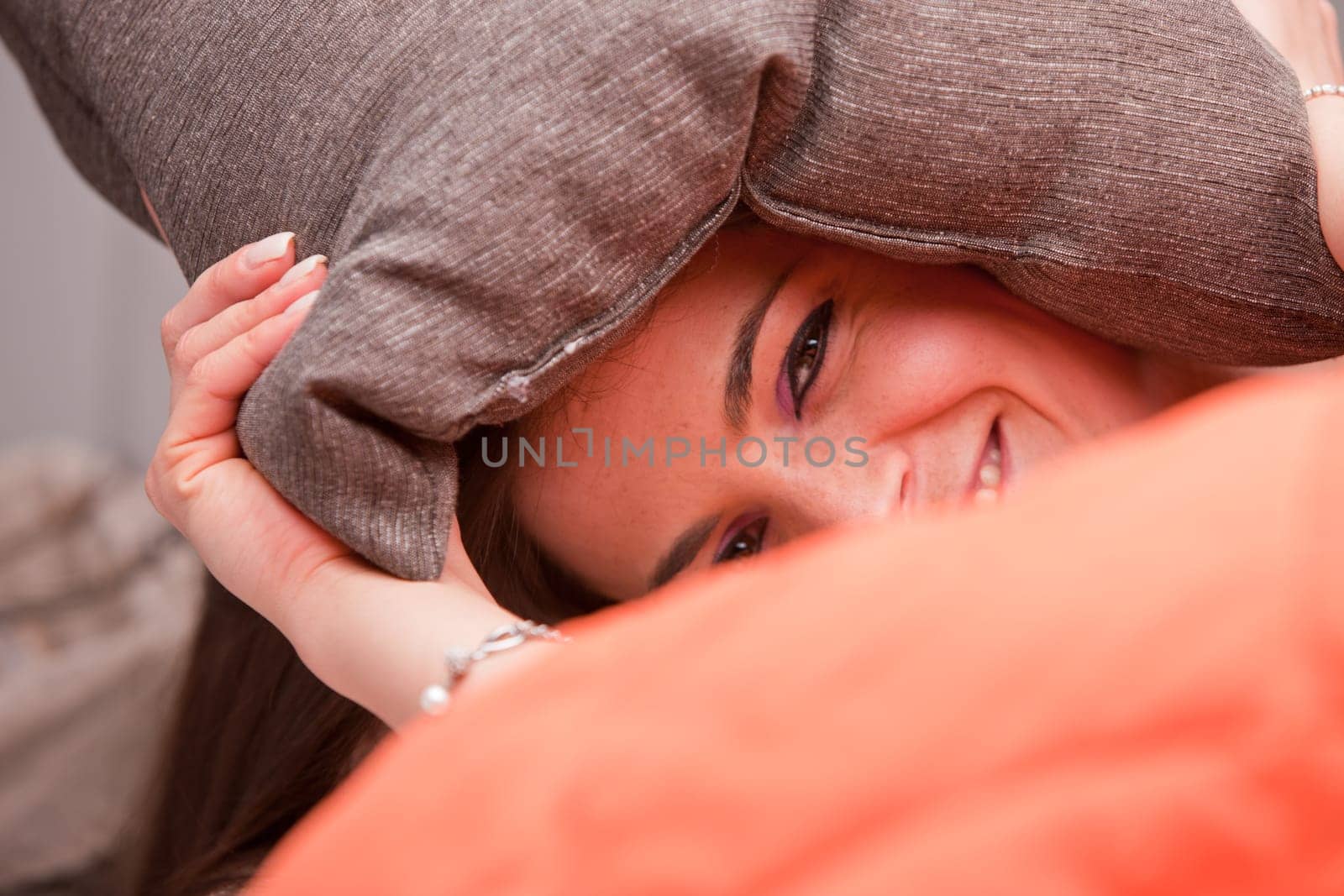 girl joking hiding behind her pillow smiling on her sofa under an orange quilt