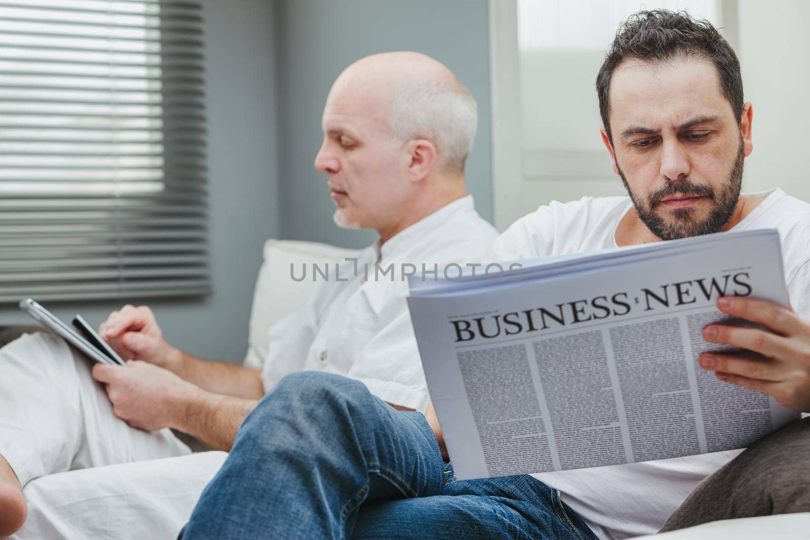 Gay couple orfather and son sitting reading the business news on a tablet and newspaoer with focus to the younger man