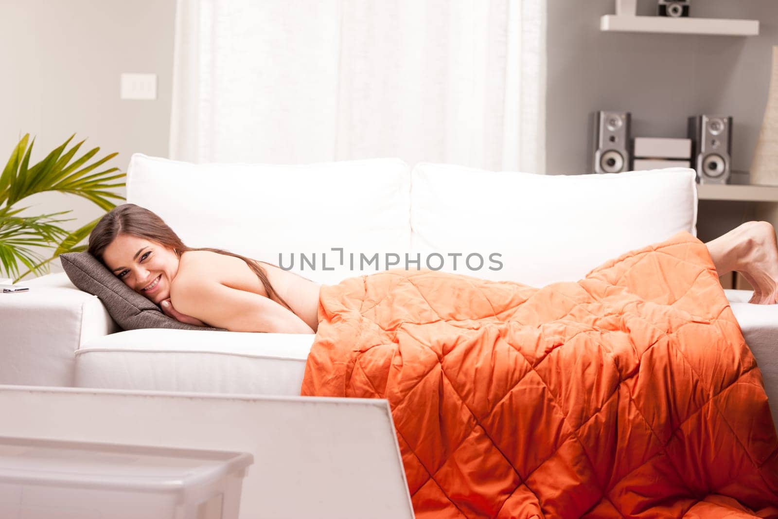 a smiling woman lying down wearing only an orange quilt but you can see only the naked skin on her back and her feet appearing