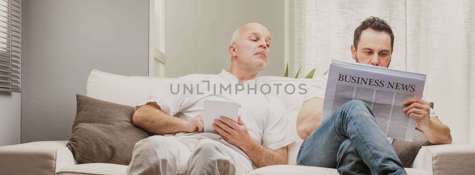 Senior man peering over the shoulder of his son or partner who is reading the Business newspaper while relaxing on a sofa