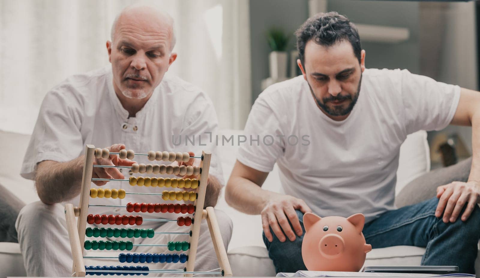 Senior man using an abacus to do financial calculations as his son sirs staring at his piggy bank