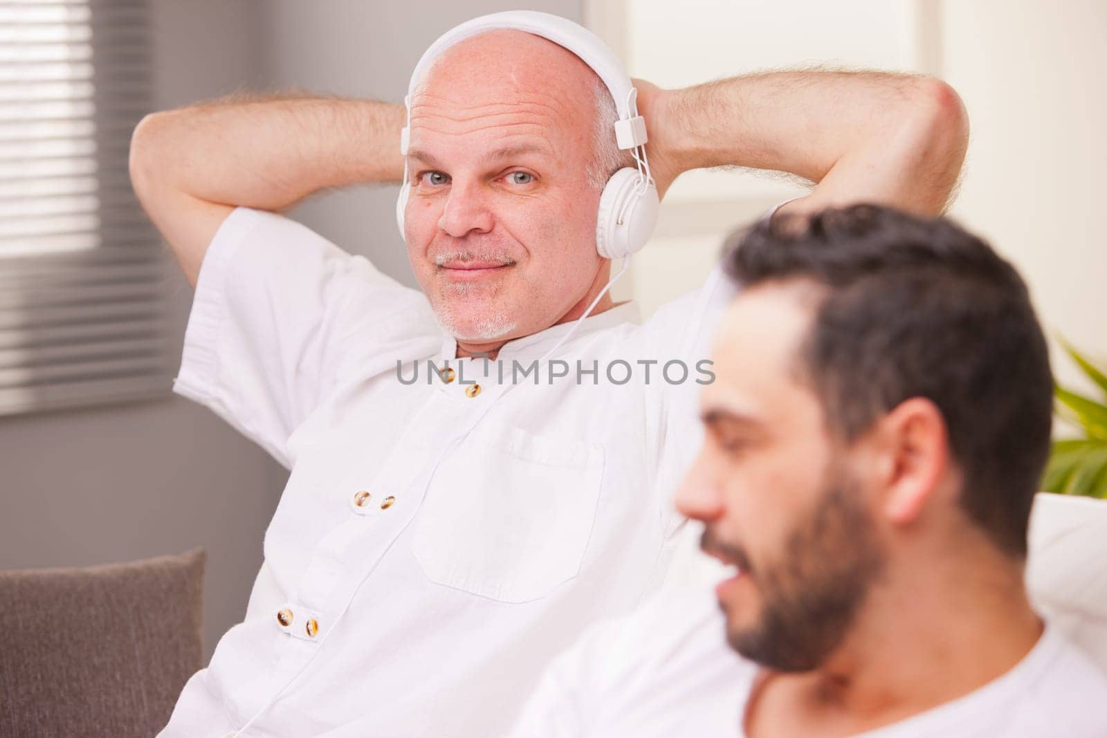 man smiling while listening to music in a living room while his friend does something else