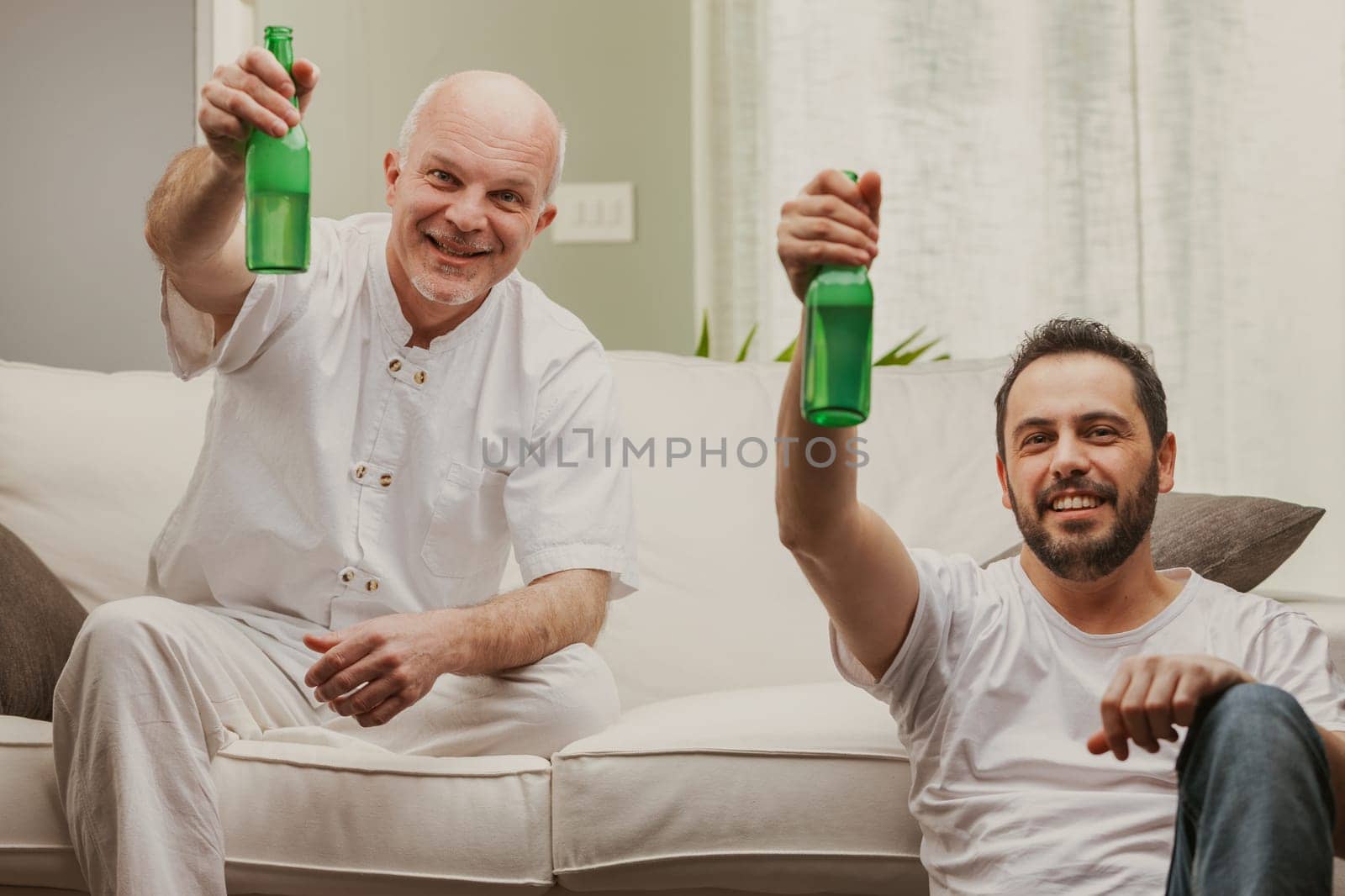 Two men celebrating toasting with bottles of beer and smiling happily at the camera as they relax on a sofa