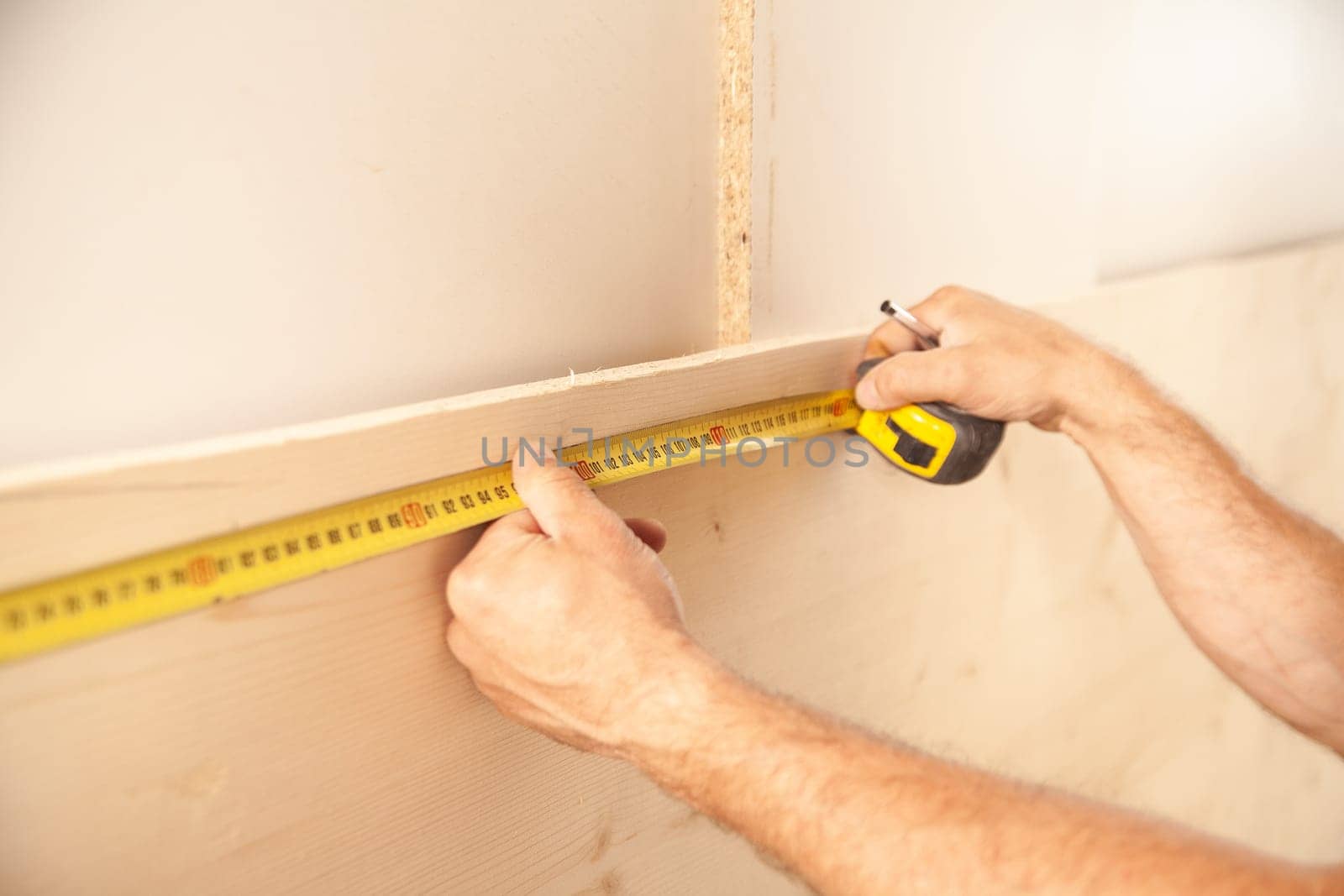 Unidentifiable man using long yellow tape measure on large wood plank leaning against white wall