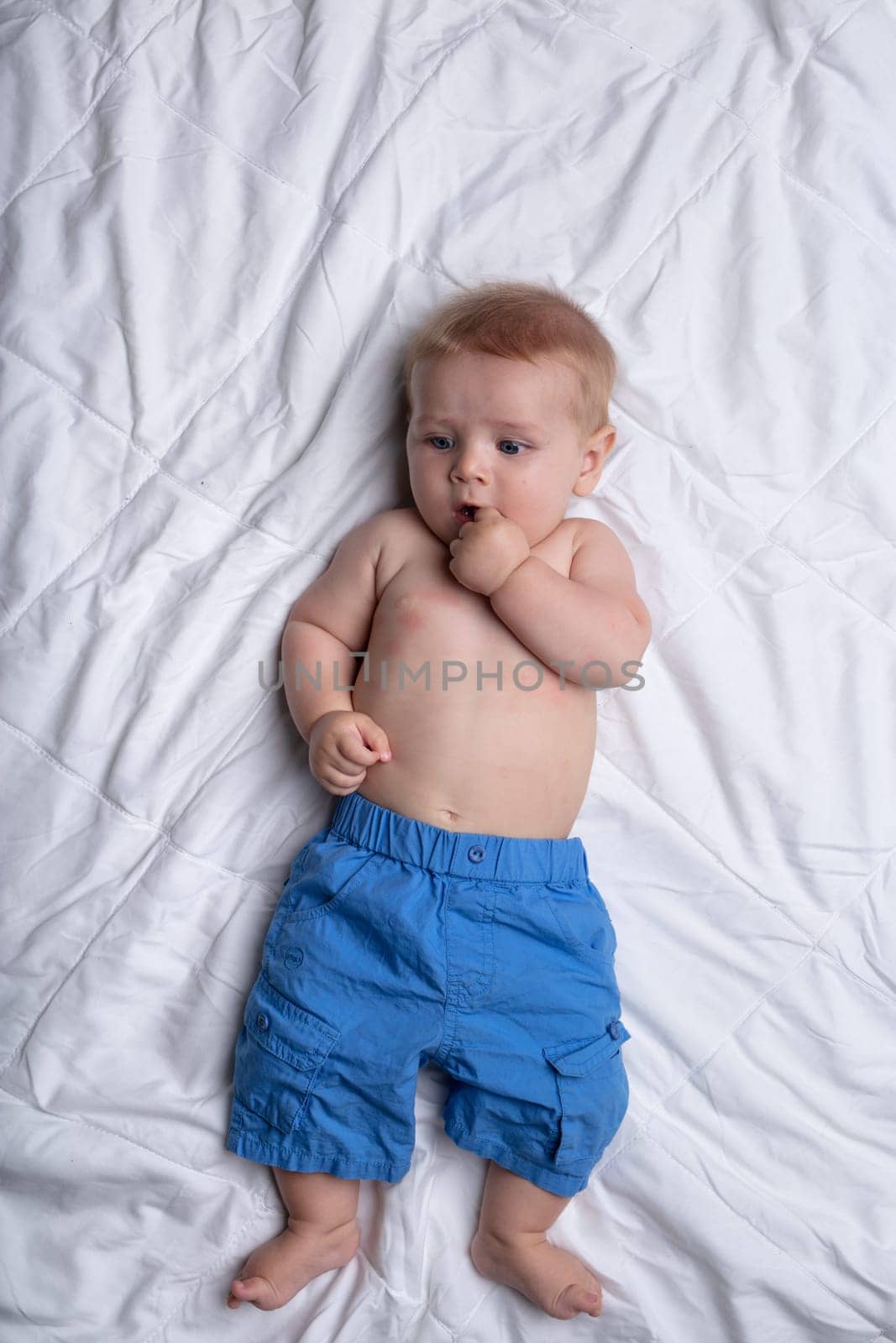 Thoughtful contented little baby boy in over sized blue pants lying on his back sucking a thumb against white bedclothes viewed from above