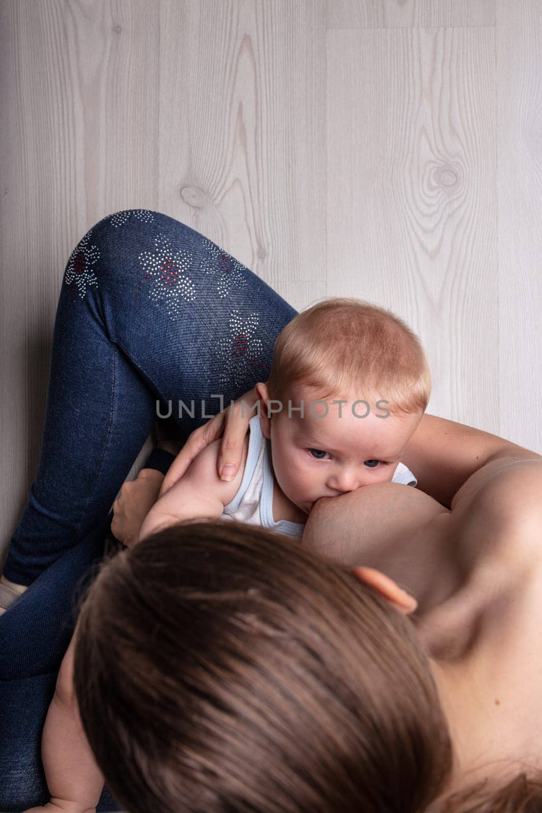 Young blond baby suckling on its mother in a view from the top of her head of the infant sucking on her teat and her bare breast, copy space on a neutral grey wood floor