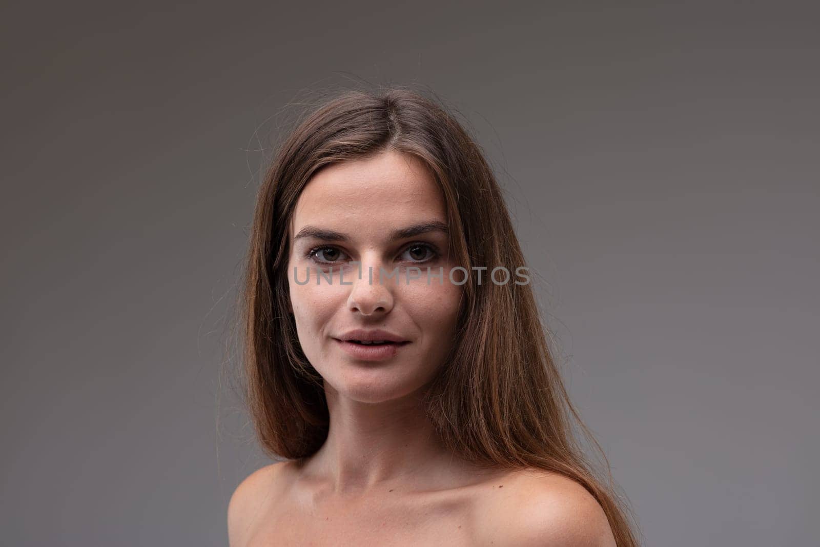 Young woman looking calmly at the camera with big brown eyes and a quiet smile as she poses with bare shoulders against a grey studio background