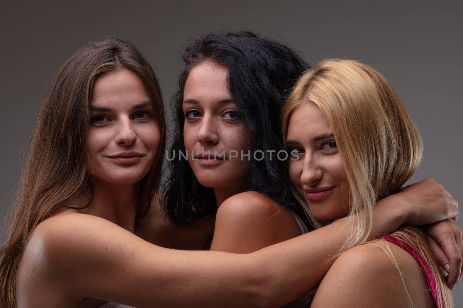 Three attractive friendly young women posing in a close embrace smiling at the camera in a head and shoulders portrait against a grey studio background