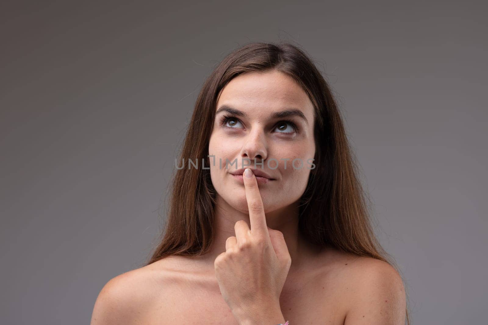 Young lady standing deep in thought with finger to lips looking up with a contemplative serious expression to blank copyspace in a head and shoulders portrait against a grey studio background