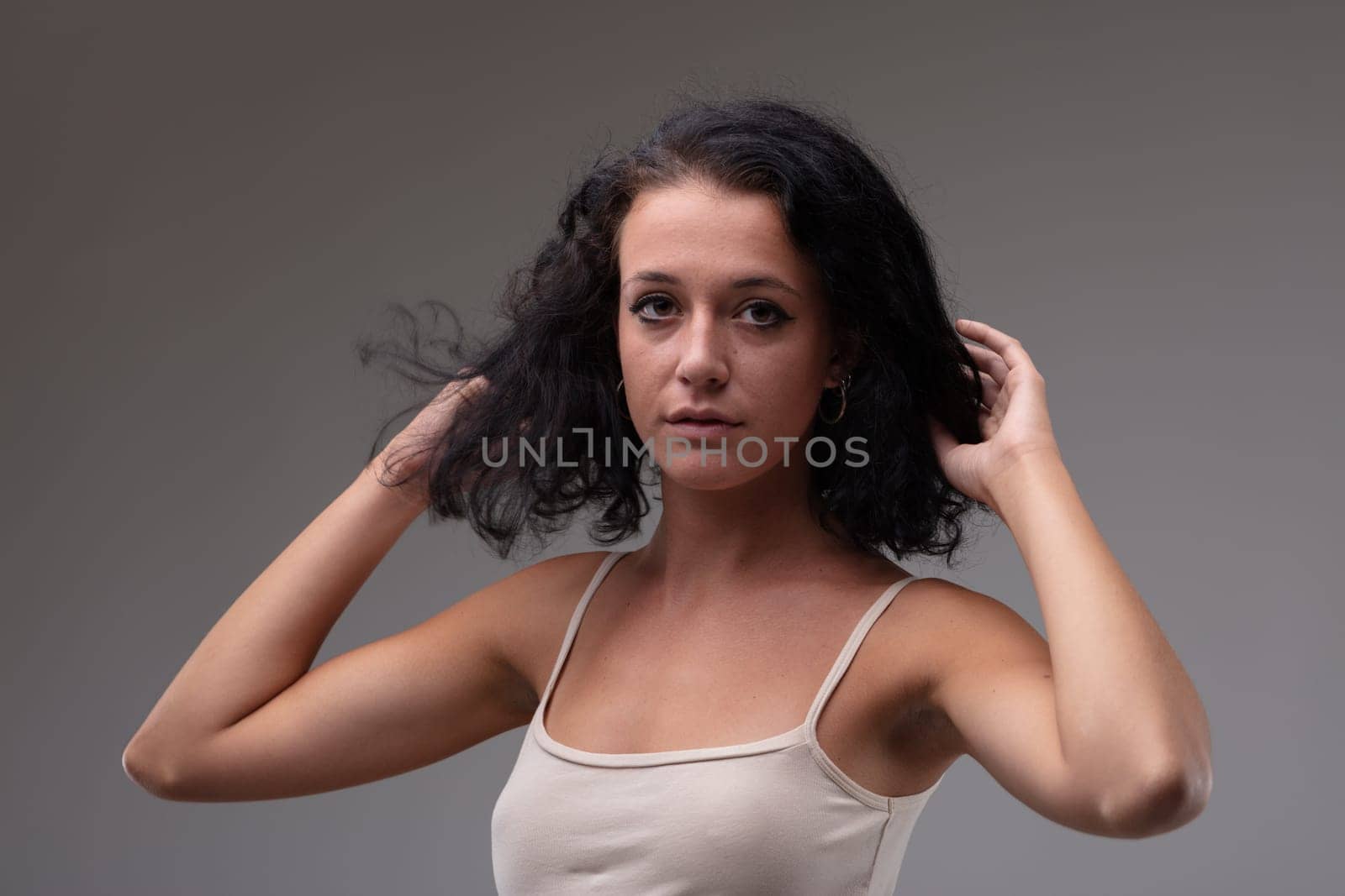 Cute young woman with curly shoulder length hair looking thoughtfully at the camera with a solemn expression and hands to head in a head and shoulders portrait against a grey studio background