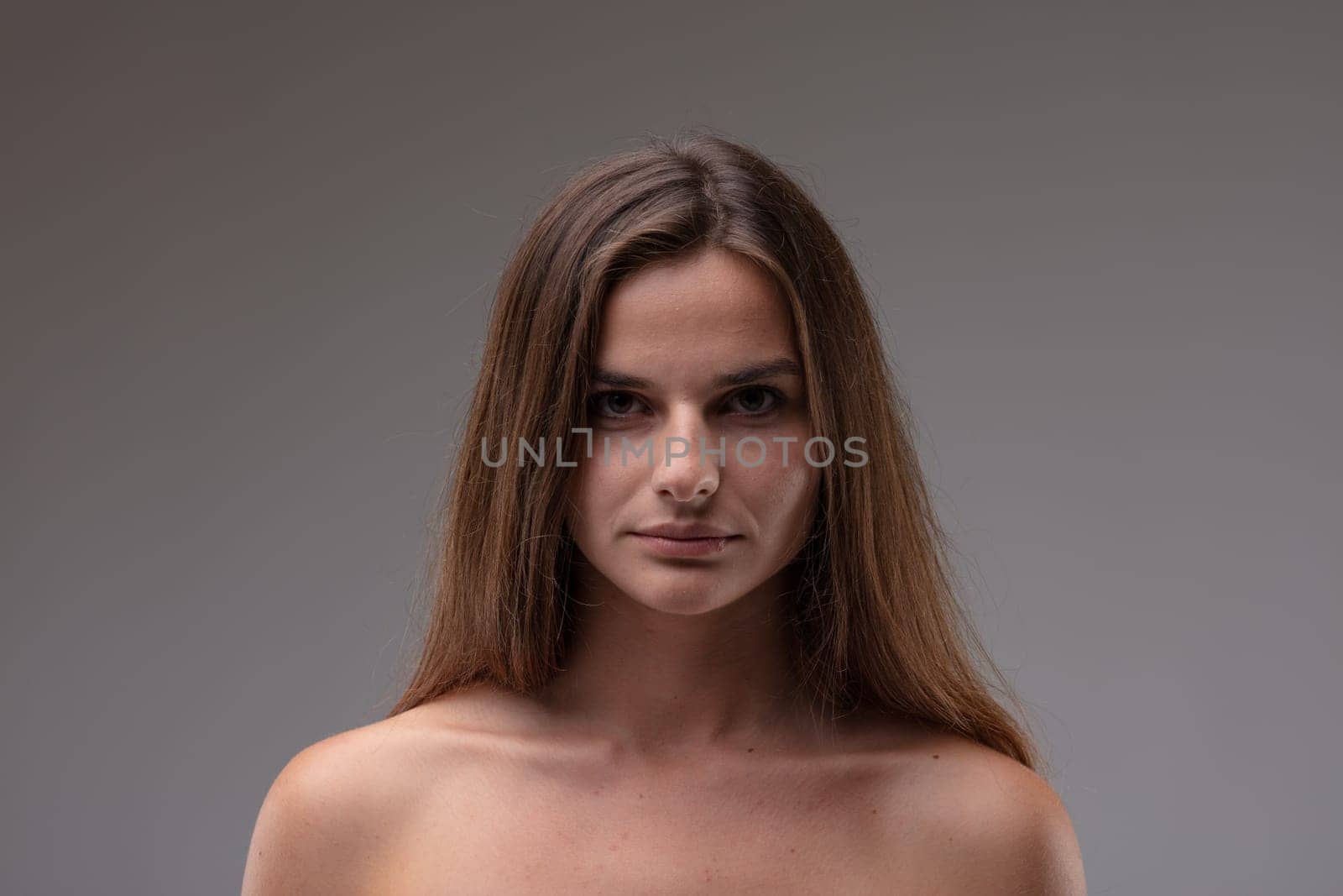 Serious attractive young brunette woman with bare shoulders staring intently at camera in a frontal head and shoulders portrait against a grey studio background