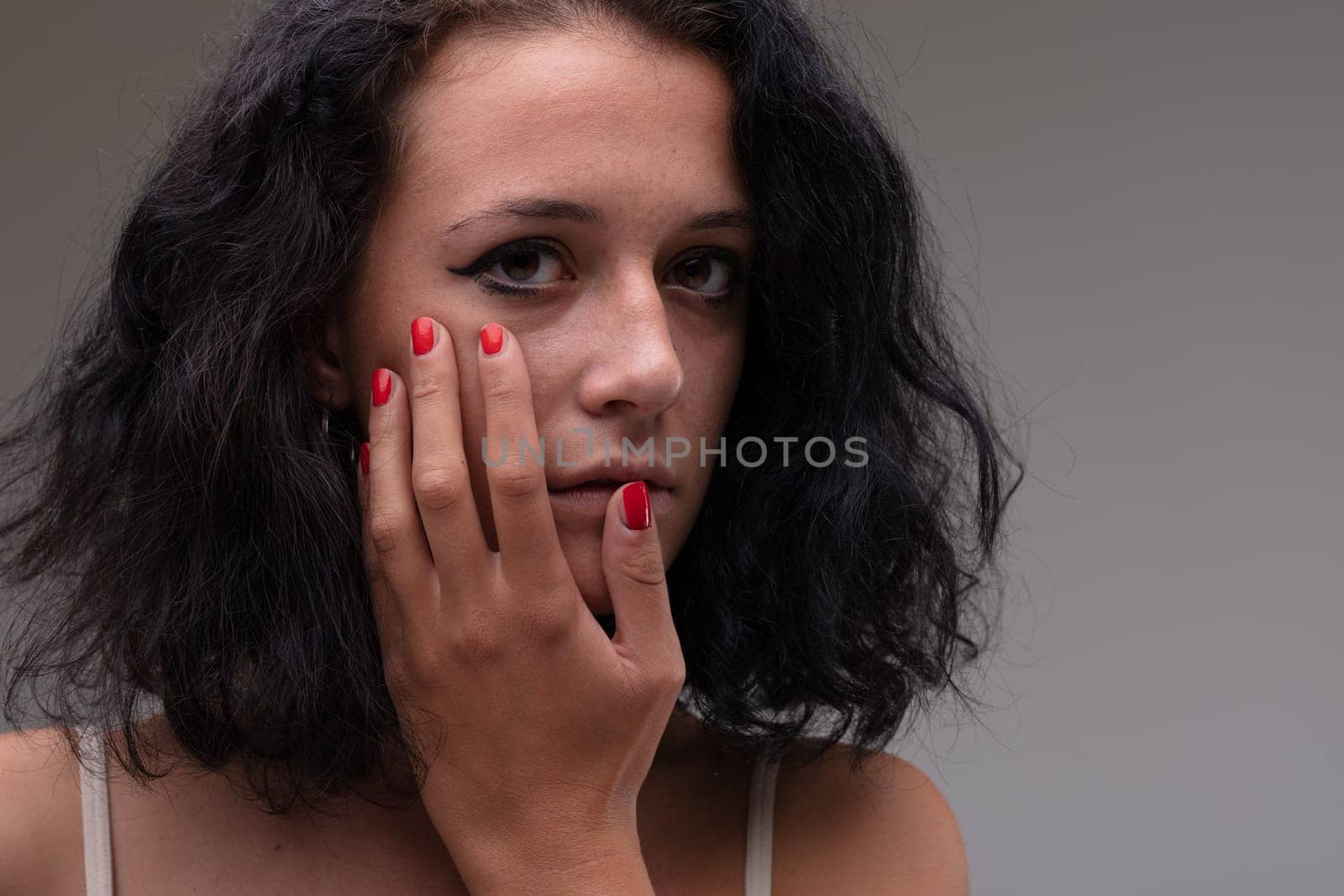 Serious soulful attractive young woman holding her hand to her cheek as she stares wide eyed at the camera with a solemn expression in a close up cropped head and shoulders portrait against a grey studio background