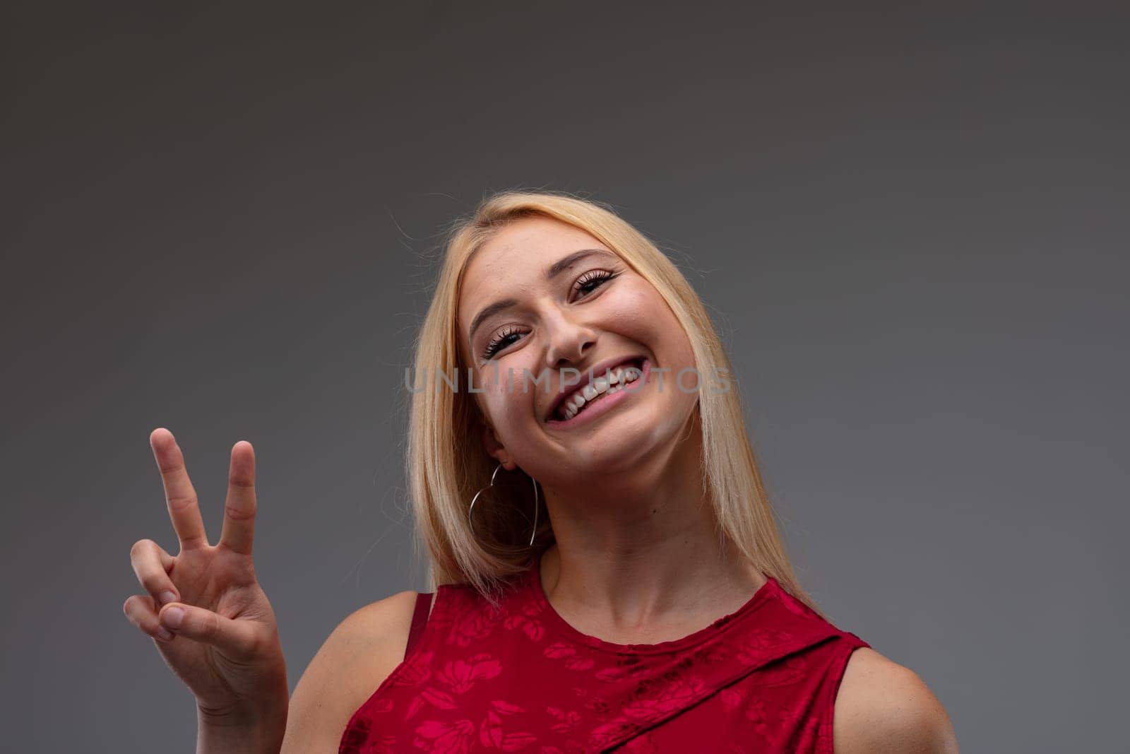 Cute charismatic blond woman in stylish red top giving a V-sign with a vivacious beaming smile of success on a grey studio background