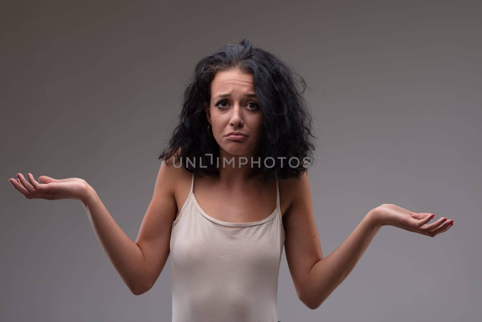 Confused young woman shrugging her shoulders with a frown of bewilderment as she gestures with her hands on a grey background