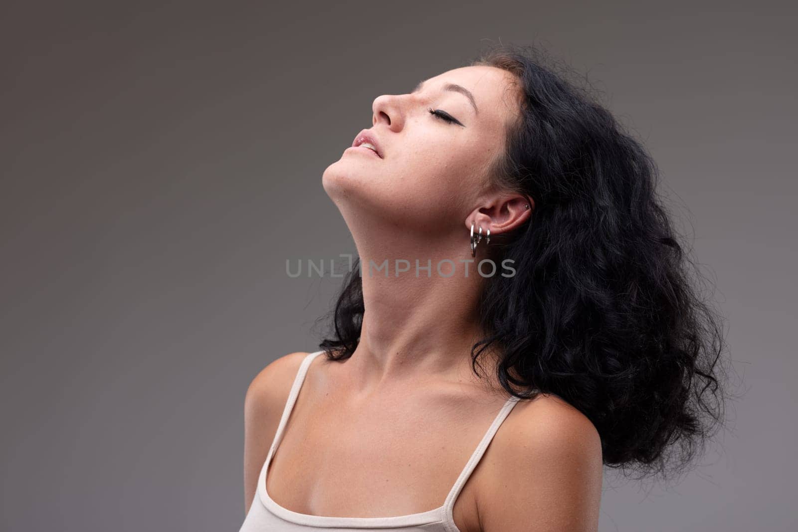 Serene young woman taking a quiet moment to relax with head tilted back, parted lips and closed eyes in a head and shoulders portrait against a grey studio background