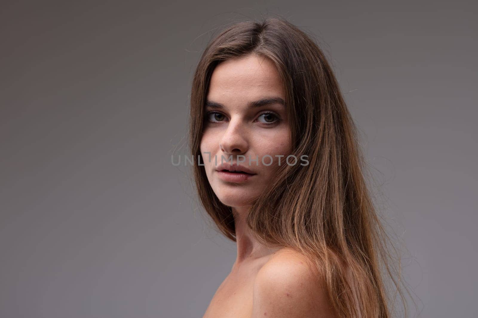 Natural portrait of a sincere young woman with naked shoulders looking back over her shoulder at the camera with wide eyes and parted lips in a head and shoulders portrait against a grey studio background