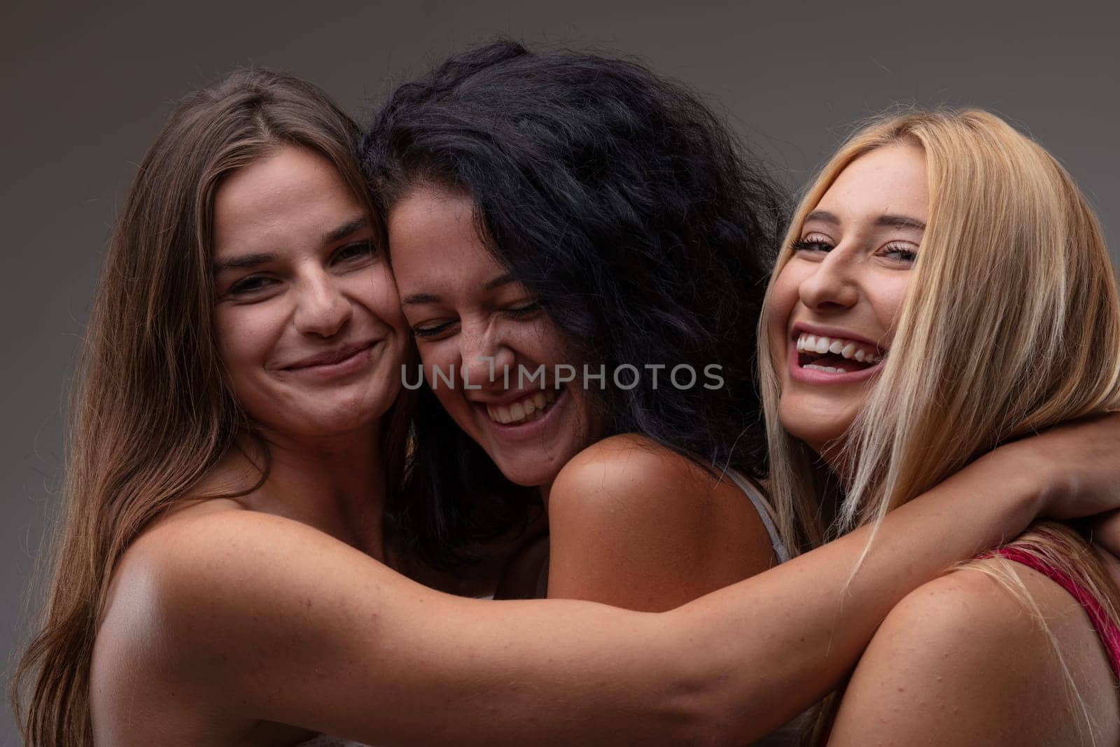 Three carefree happy young woman laughing at the camera as they pose arm in arm in a close embrace in a head and shoulders portrait against a grey studio background