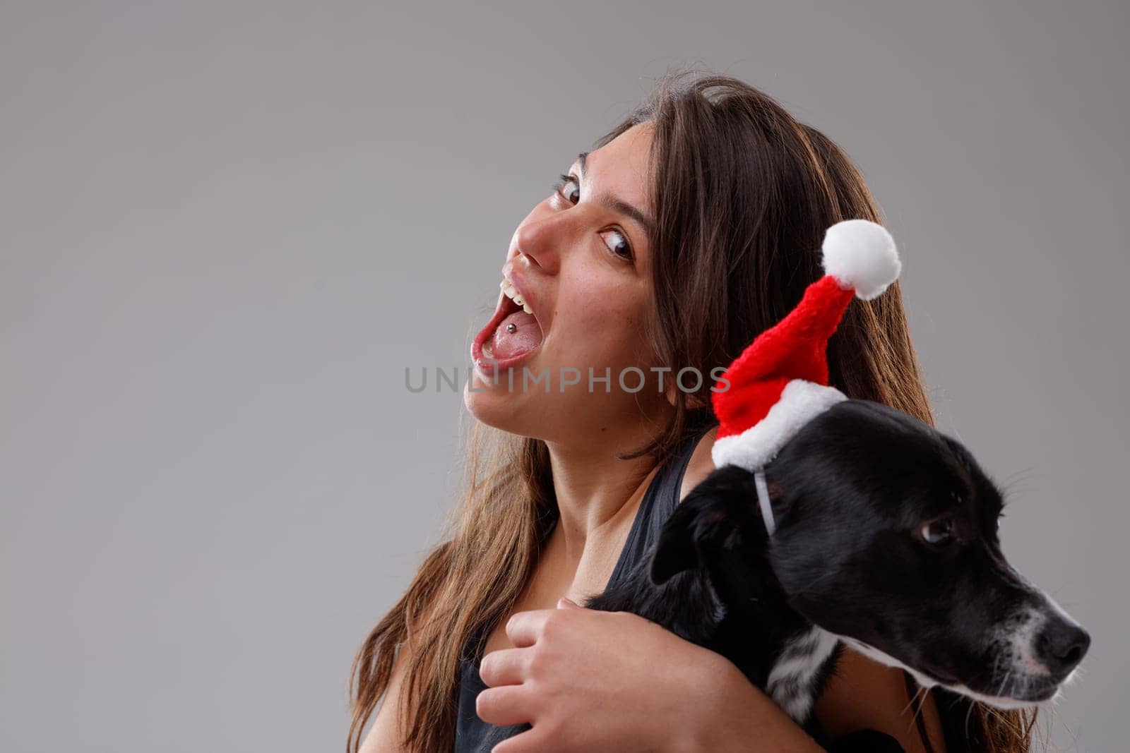 Young woman and her pet dog celebrating Christmas in festive red outfits as she hugs it in her arms while displaying the piercing in her tongue to the viewer