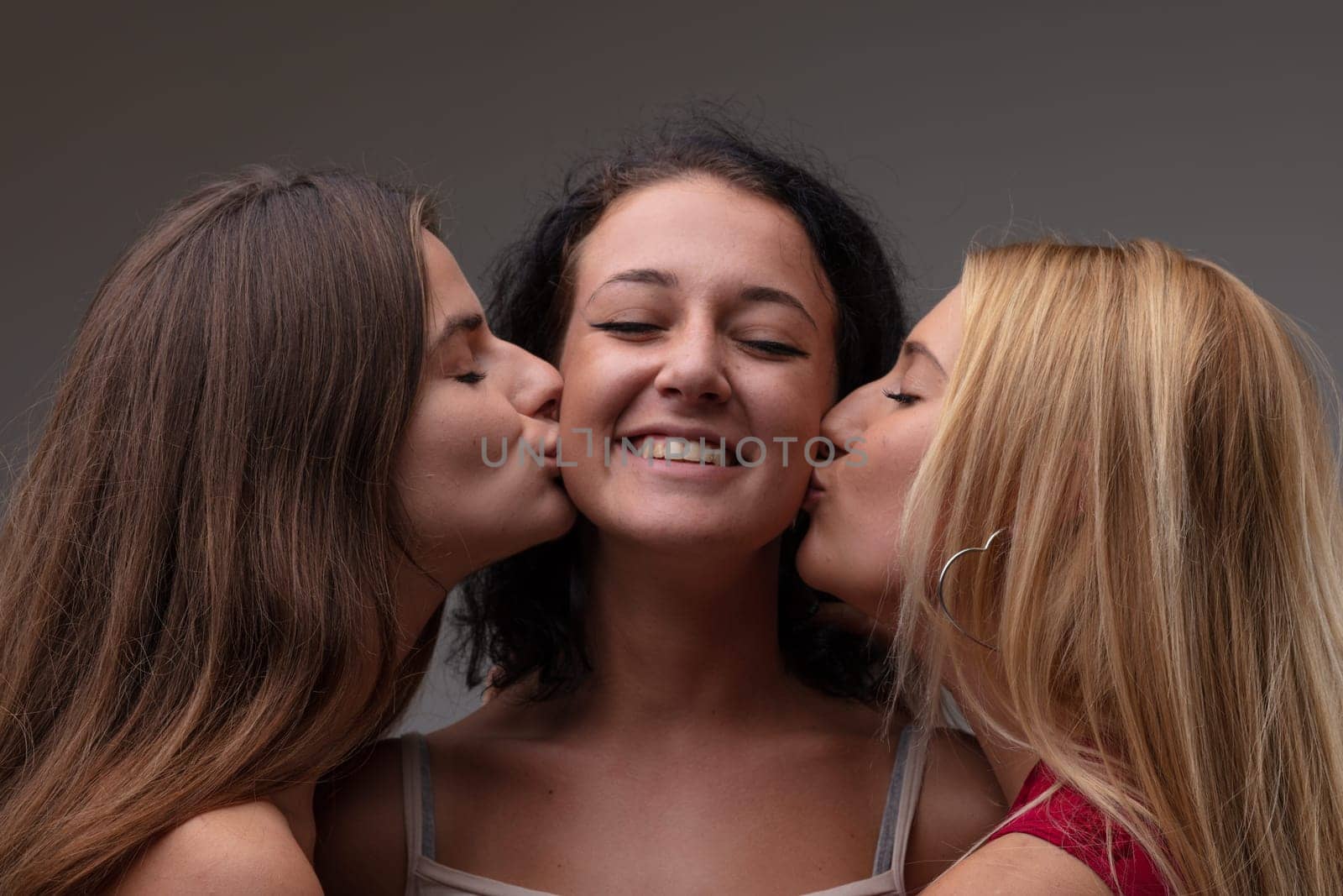 Happy young woman with a lovely warm smile of pleasure being kissed on the cheeks by two of her friends in a head and shoulders portrait against a grey studio background