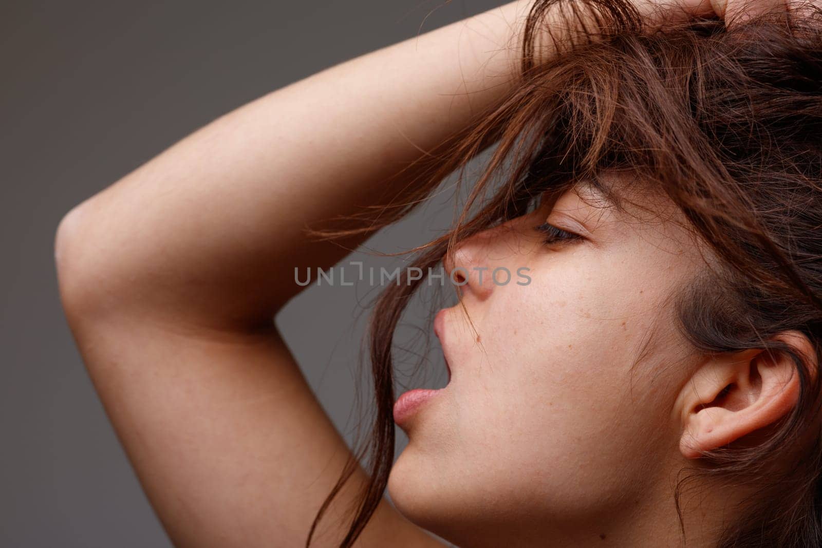 Sensual cropped portrait of a provocative young woman posing with mouth open and hand raised to her tousled long brown hair viewed in profile