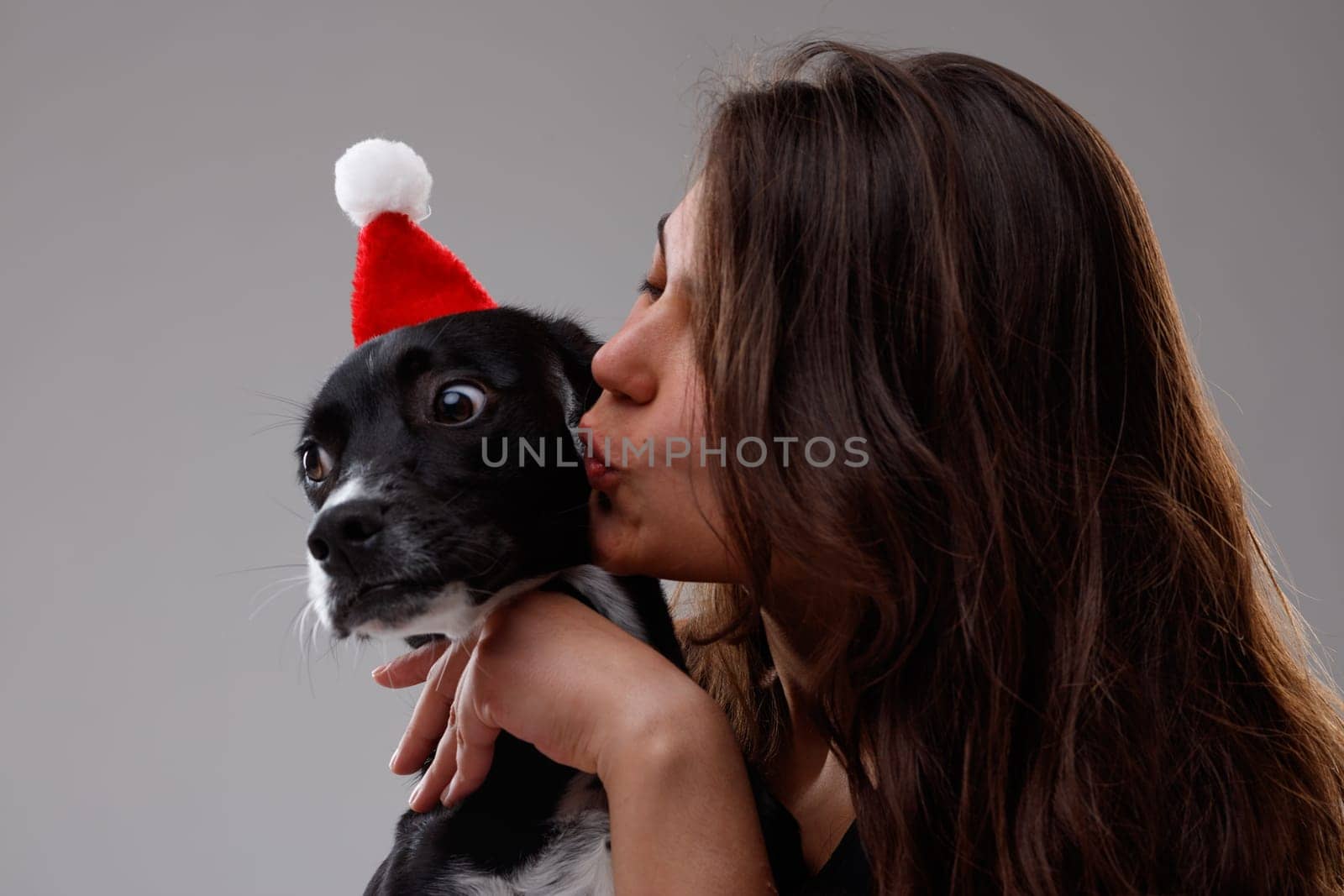 Loving young woman kissing her little pet dog wearing a festive red Santa hat for Christmas as she hugs it in her arms
