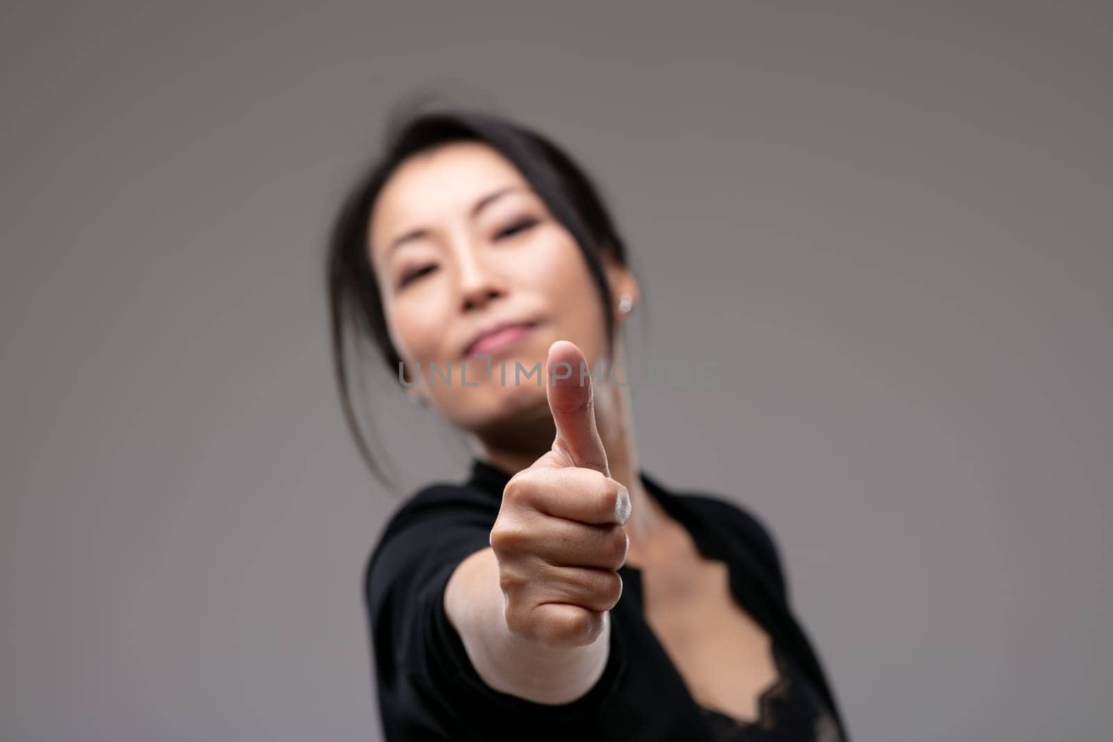 Pleased confident Asian woman giving a thumbs up gesture of success, approval and agreement with focus to her hand over a grey studio background